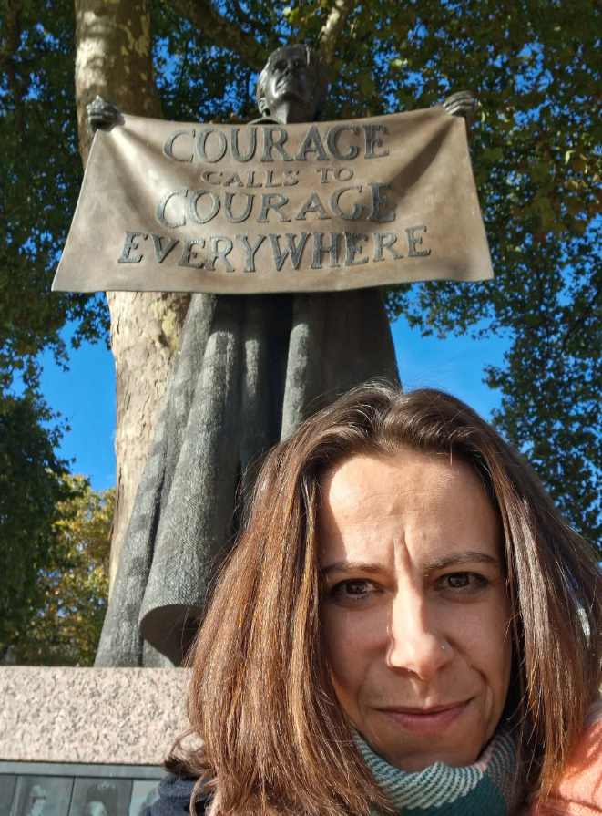 A woman taking a selfie in front of a statue of a person holding a banner that reads 'Courage Calls to Courage Everywhere,' with trees and a blue sky in the background.