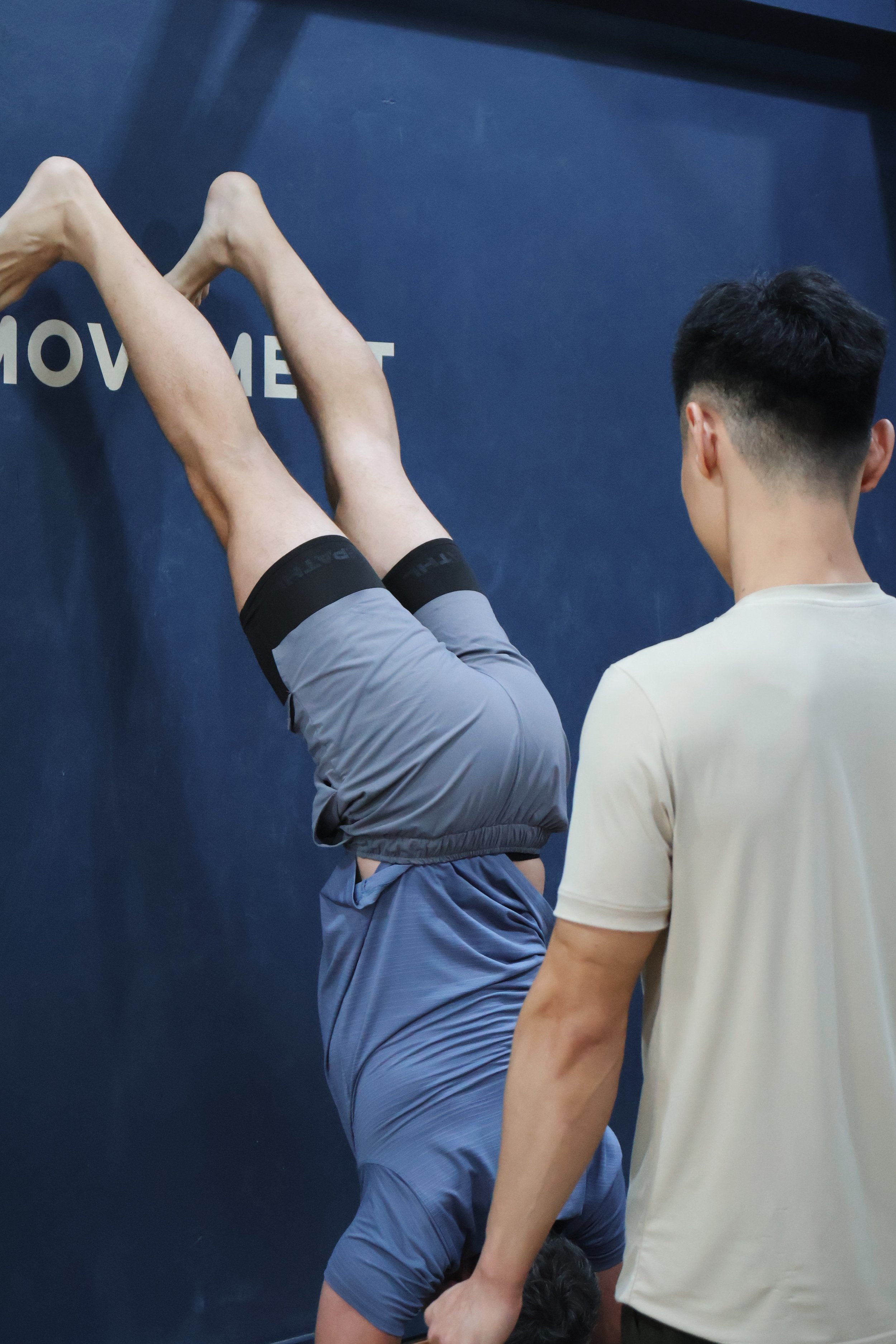 Two young men are performing a wall handstand, facing the wall with their hands on the floor and feet against the wall. A third person observes them.