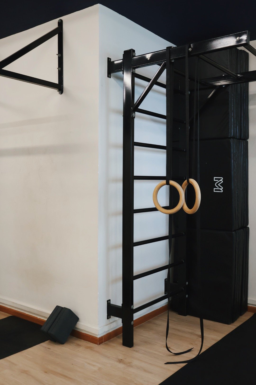 Home gym corner with black wall-mounted pull-up rack, gymnastic rings hanging from black straps, stacked black padded mats, black weight plates or storage blocks, and a black cushioned exercise mat on light wooden floor.