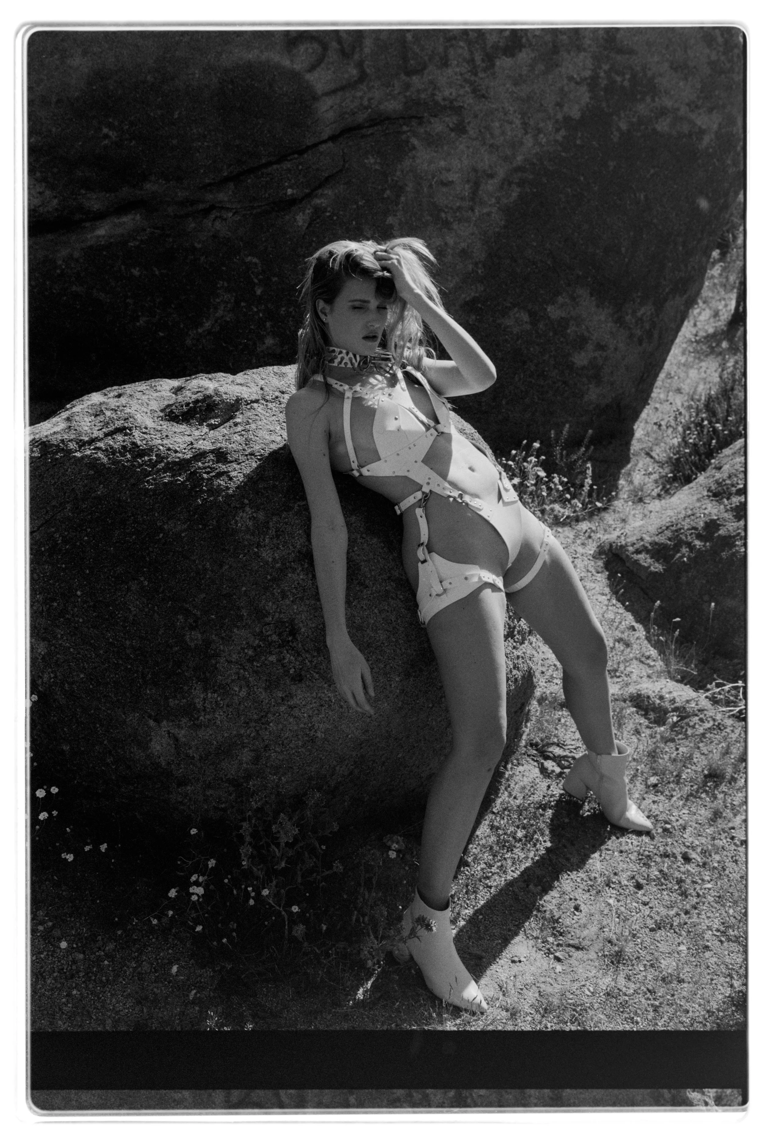 A woman with long hair, wearing a harness and boots, posing on large rocks in a natural outdoor setting. The photo is in black and white.
