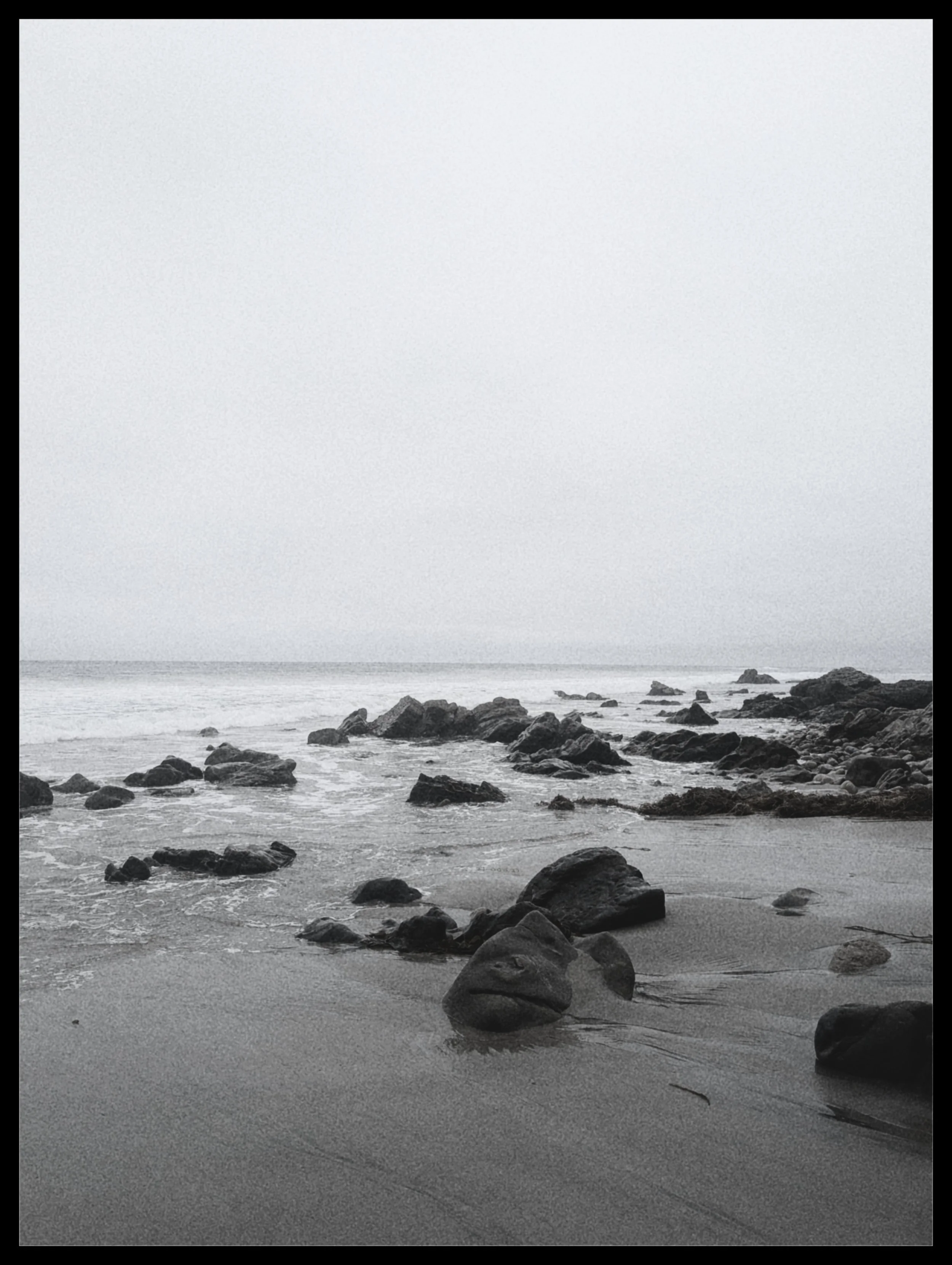 Black and white photograph of a rocky shoreline with water and sand, under an overcast sky.