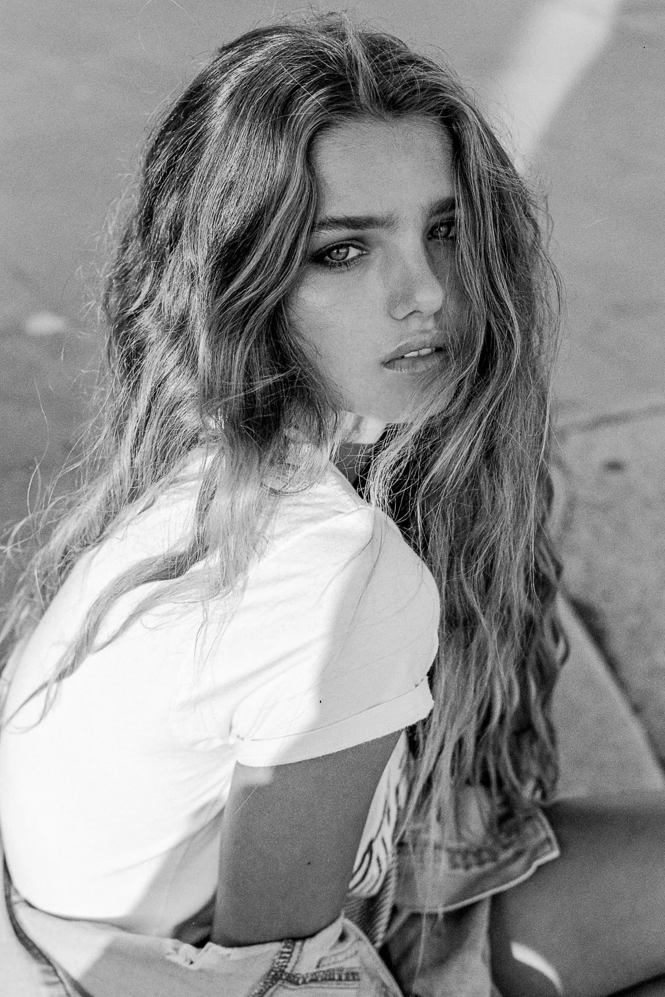 Black and white photograph of a young woman with long, wavy hair looking at the camera with a serious expression, sitting outdoors with a blurred background.