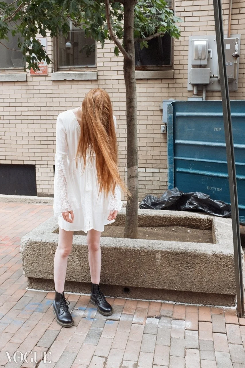 A young woman with long red hair wearing a white dress and black combat boots standing outdoors in front of a tree and a brick building. She is looking down with her head bowed, and her hands at her sides.