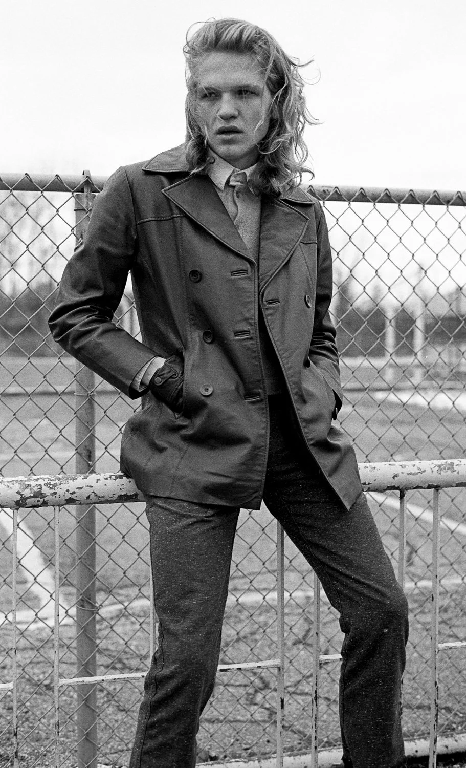 Black and white photo of a woman with shoulder-length wavy hair wearing a trench coat, standing in front of a chain-link fence, looking at the camera with a serious expression.