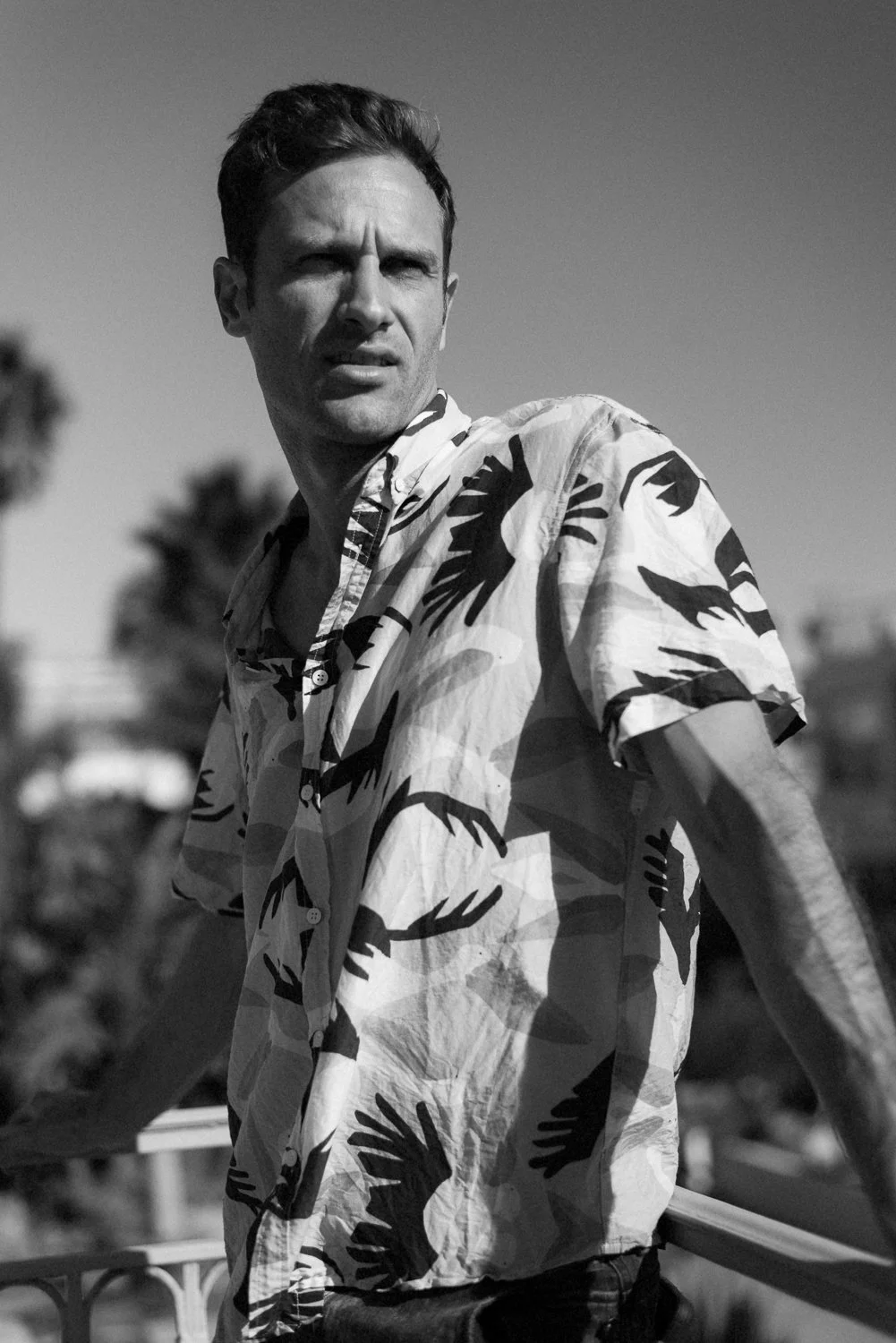 A man with short dark hair, wearing a patterned short-sleeve shirt, looks to his right outdoors with a background of palm trees and cloudy sky.