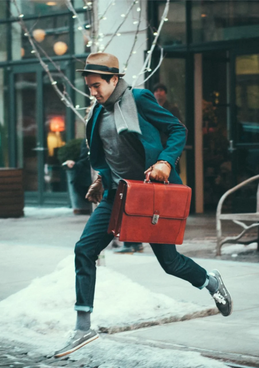 A young man in a suit with a scarf and fedora hat running on a city sidewalk, carrying a red briefcase.