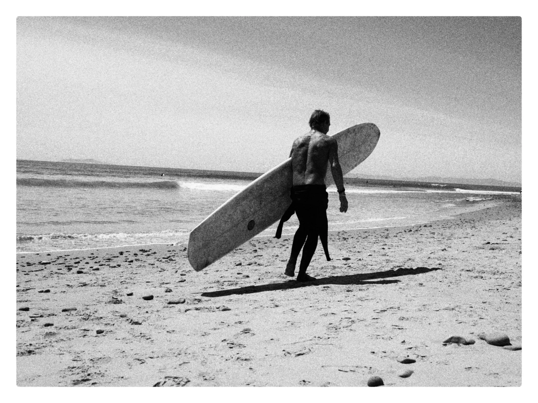 A person walks along the beach holding a surfboard, with rolling waves and a cloudy sky in the background, in a black-and-white photo.