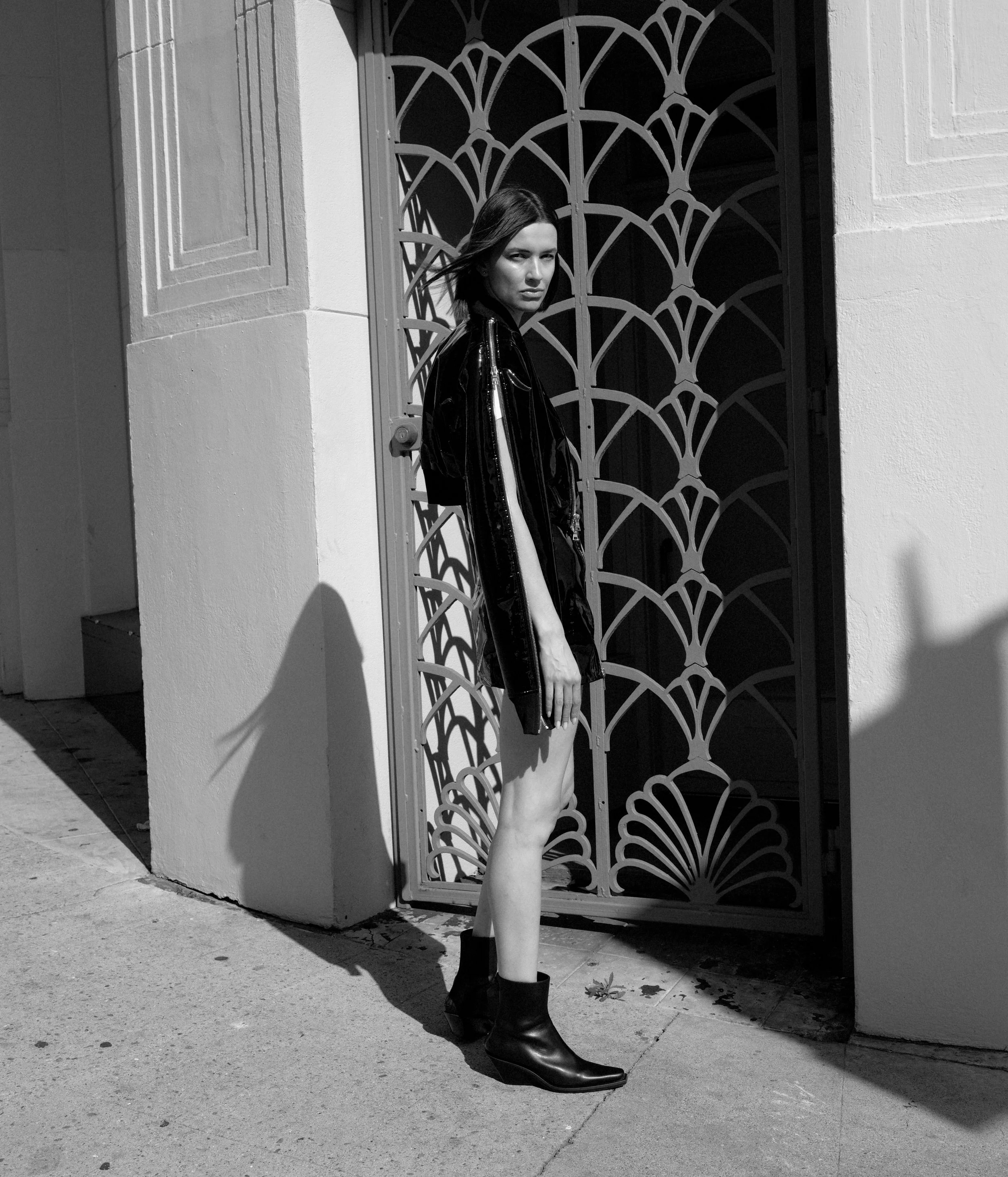 A woman wearing a leather jacket and black boots standing outdoors next to a decorative metal gate, with shadows cast on the ground in sunlight.