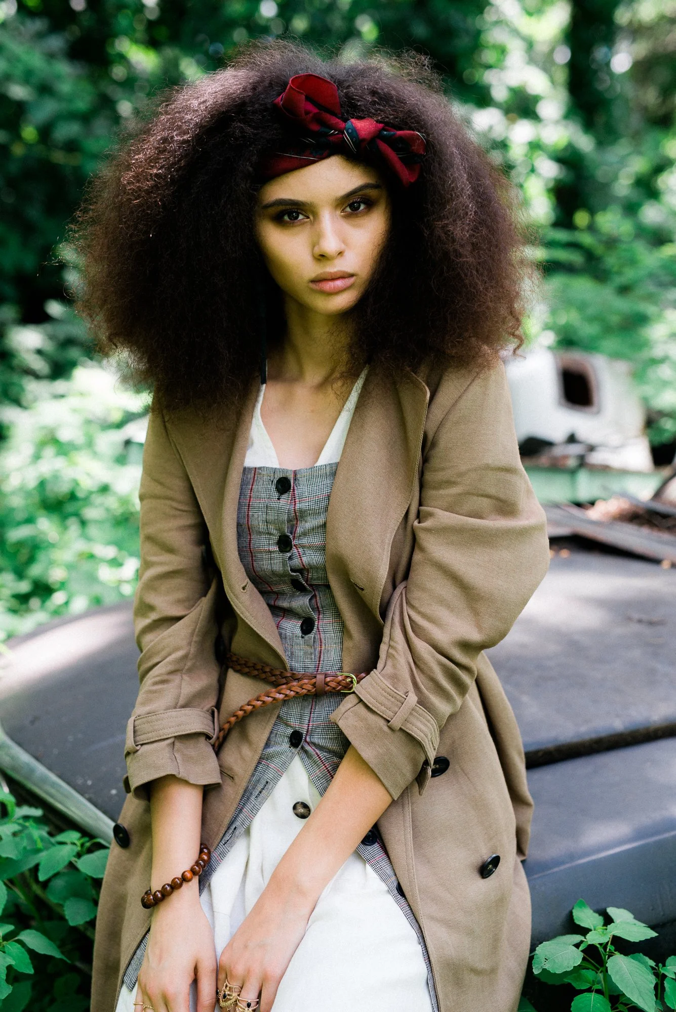 A young woman with voluminous curly hair wearing a red headscarf tied around her head, outdoors surrounded by greenery, dressed in a beige trench coat over a plaid dress, with jewelry on her hands and wrist.