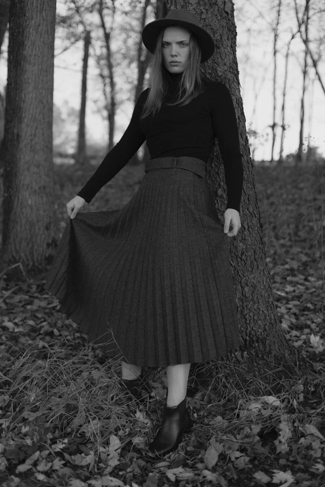 A woman standing by a tree in a wooded area, wearing a black long-sleeve top, a wide-brimmed hat, a long pleated skirt, and ankle boots.