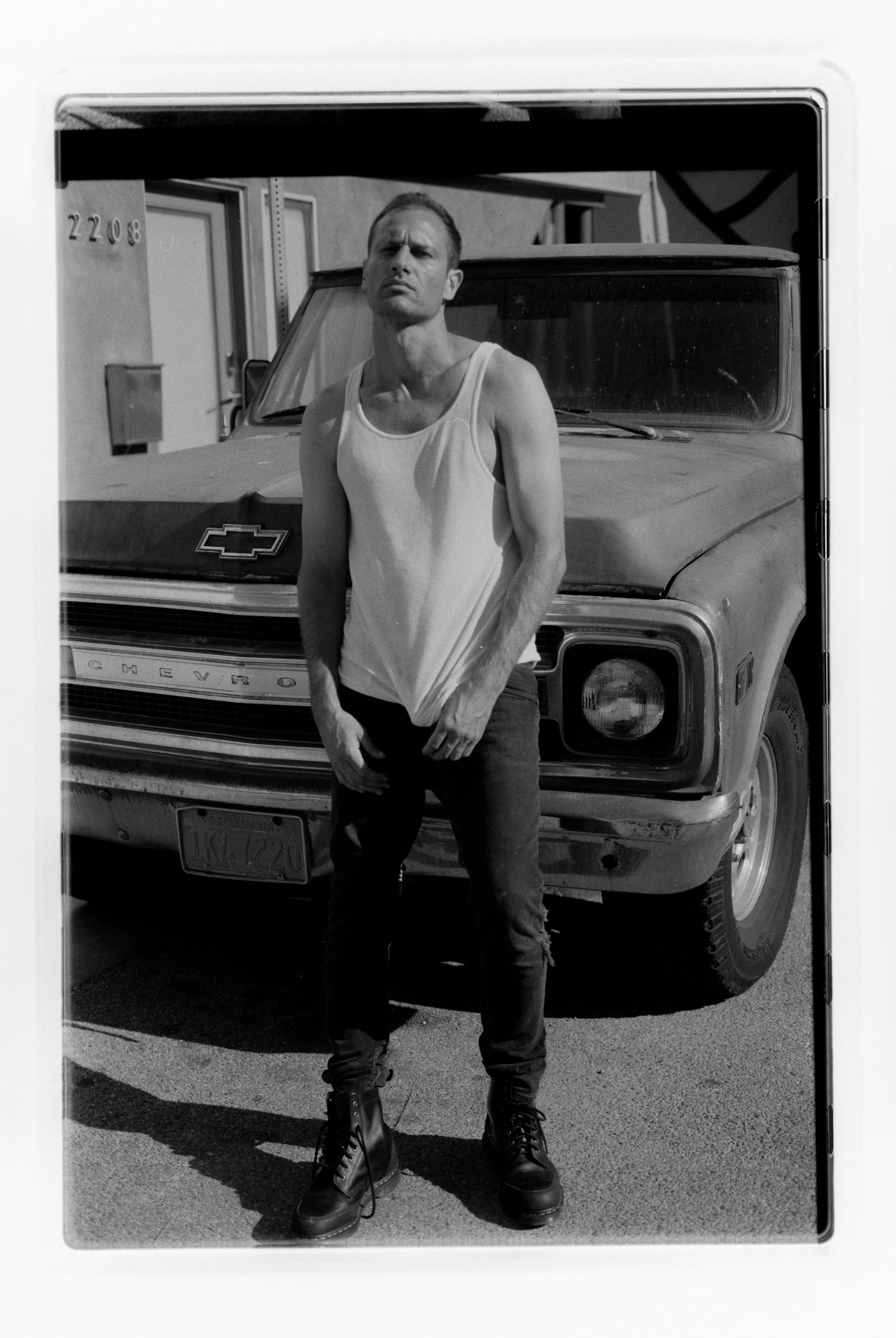 Black and white photo of a man standing in front of an old Chevrolet pickup truck, wearing a sleeveless shirt and combat boots, looking directly at the camera with a serious expression.