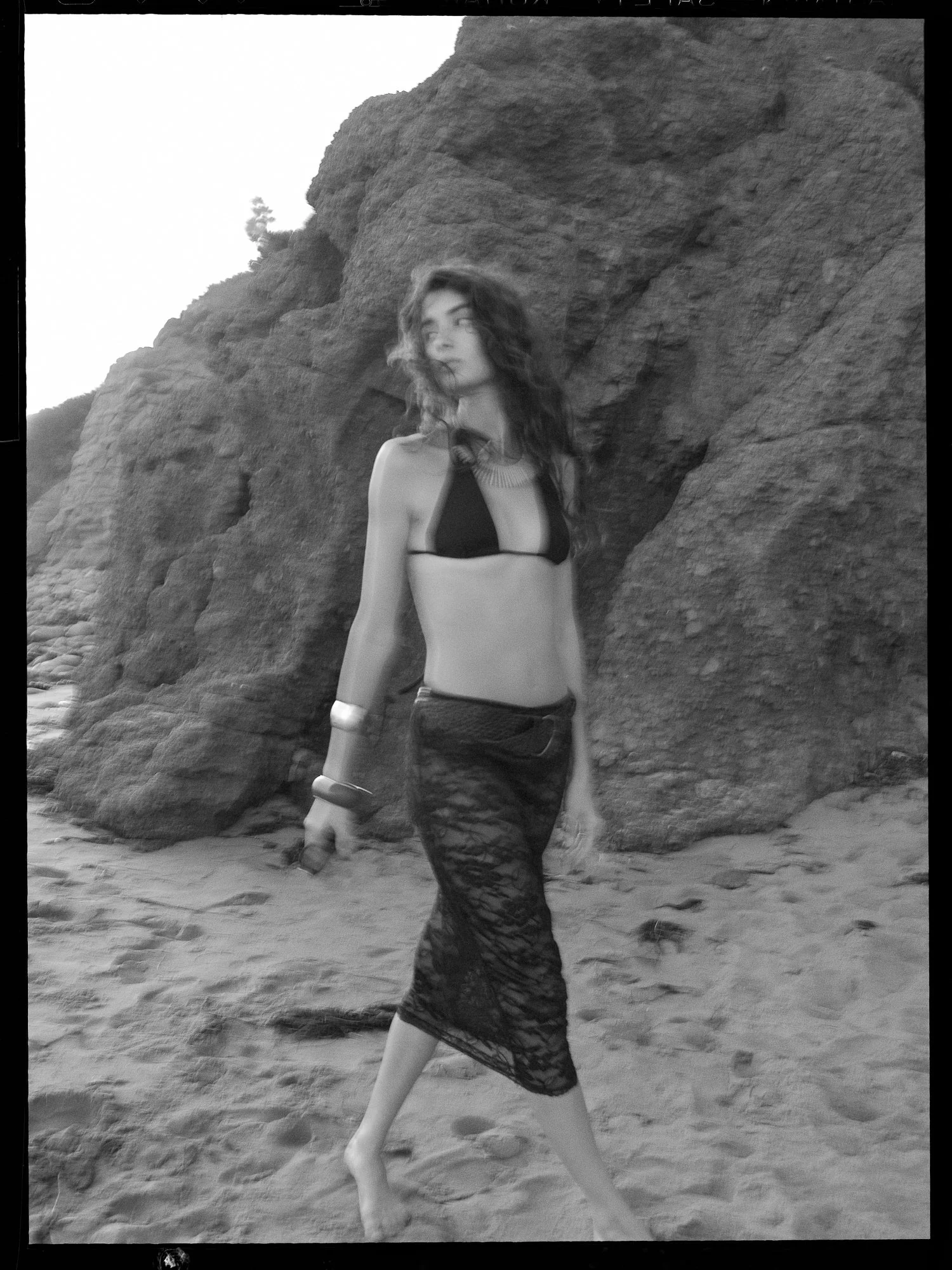 A woman walking on a sandy beach next to large rocky formations, wearing a sleeveless top and a lace skirt.