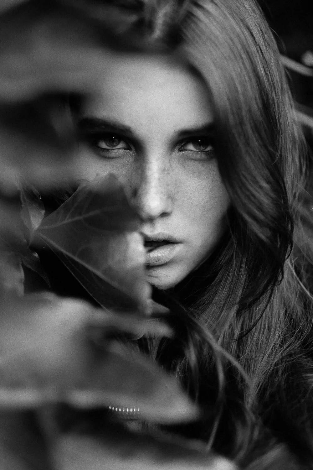 A black and white close-up of a woman with intense eyes, partially obscured by leaves.