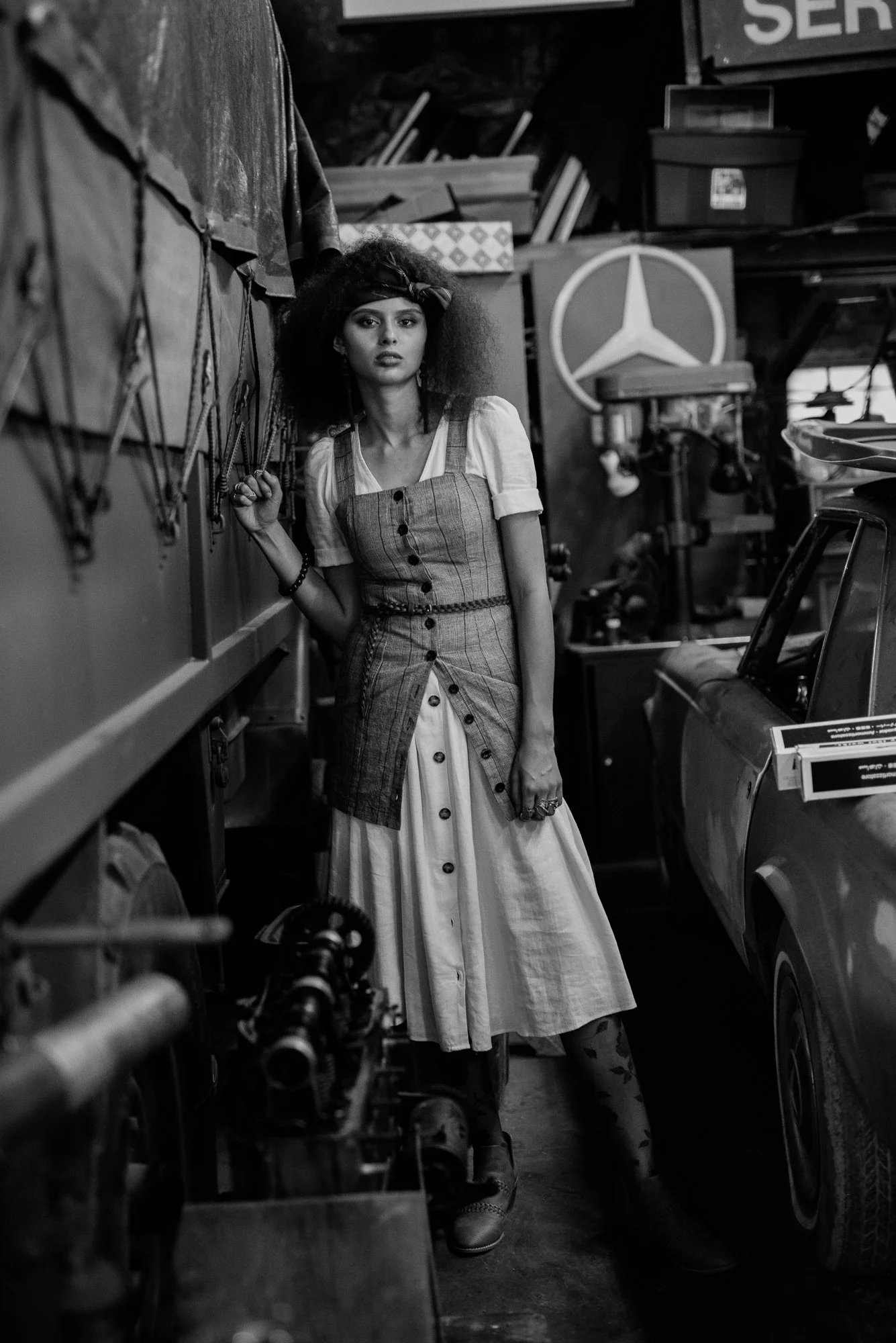 A woman with curly hair and a headband stands in a cluttered workshop among various tools and industrial equipment, wearing a plaid apron over a dress.