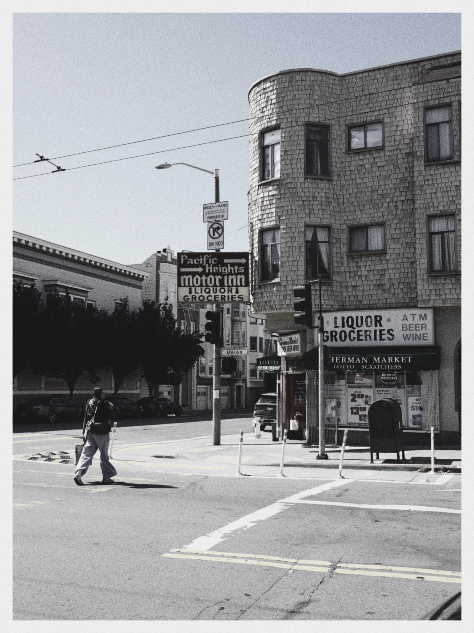 A black-and-white street scene featuring an intersection in front of a liquor and grocery store with signs for beer, wine, lotto, and scratchers. A man walks across the street, and a woman stands nearby. There are parked cars and trees along the stre