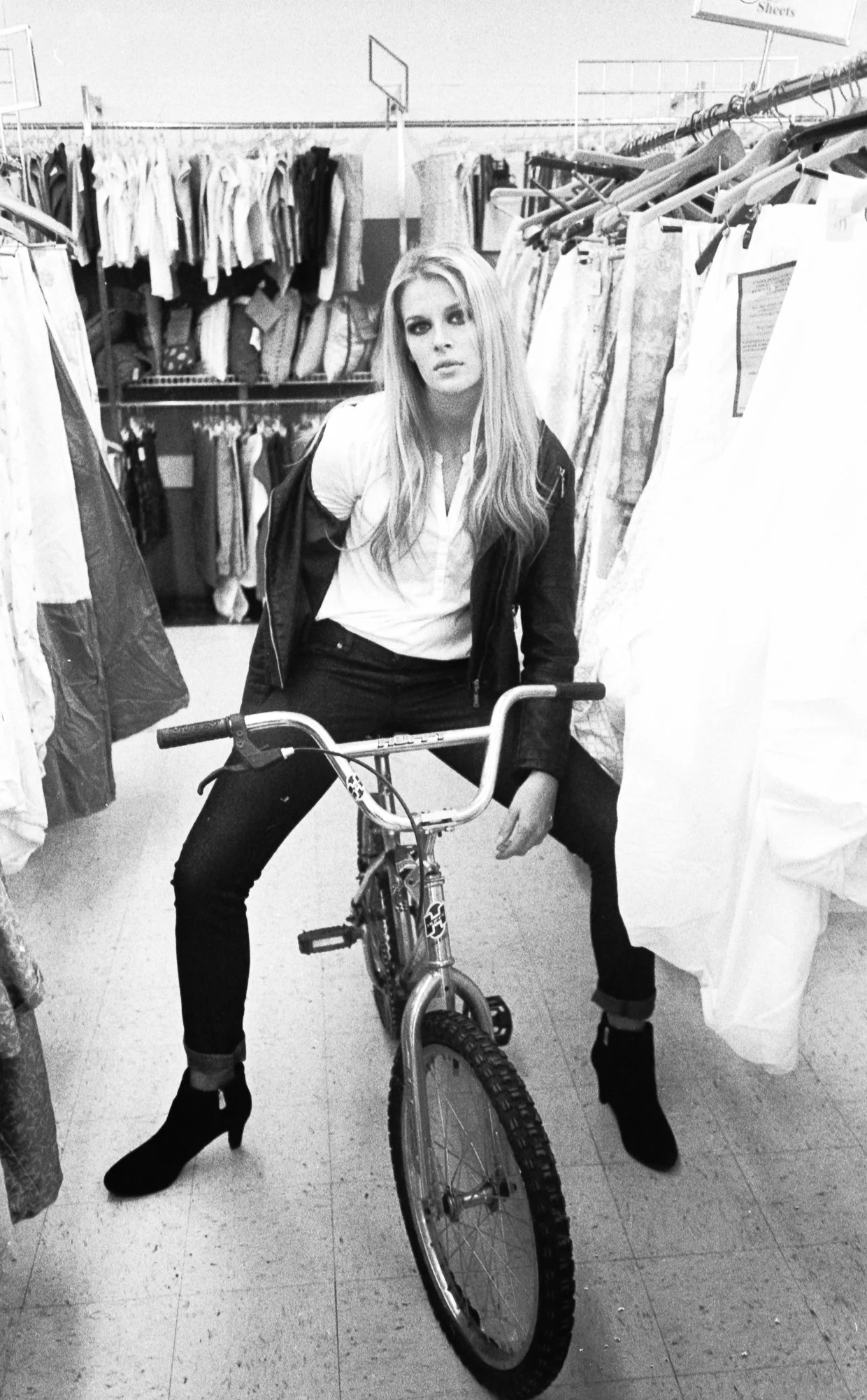 A woman with long blonde hair wearing a white shirt, black jacket, and black high-heeled boots sitting on a bicycle in a store aisle with racks of clothing on both sides.