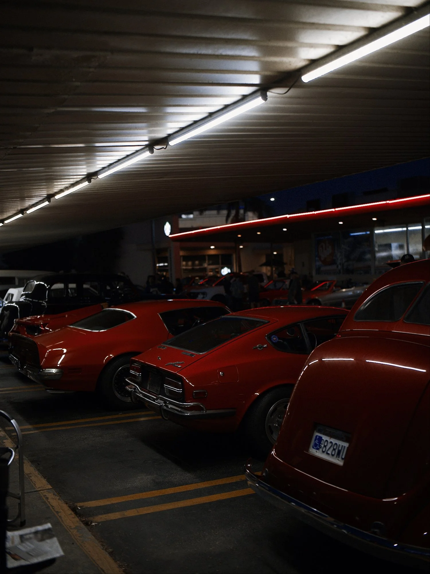 Dark parking garage with classic red cars parked in specific spots, illuminated by fluorescent lights overhead, with city buildings visible outside through large windows.
