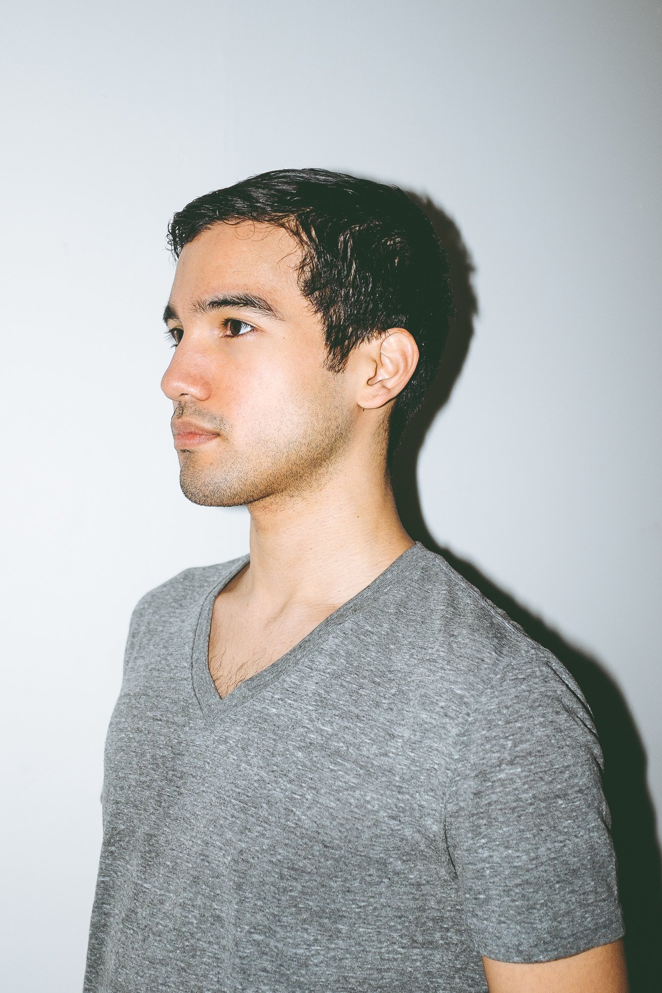 Side profile of a young man with dark, wavy hair and light skin, wearing a gray V-neck T-shirt, standing against a plain white background.