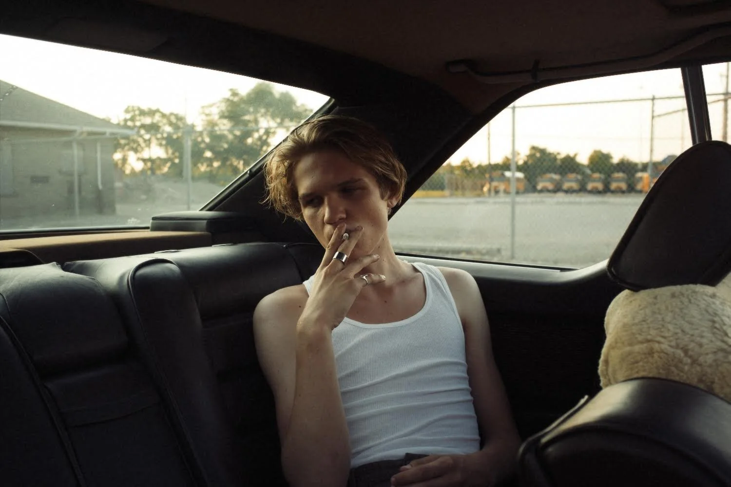 A young man with wavy hair wearing a white tank top sitting in the backseat of a car, smoking a cigarette, with a sunset or evening light outside.