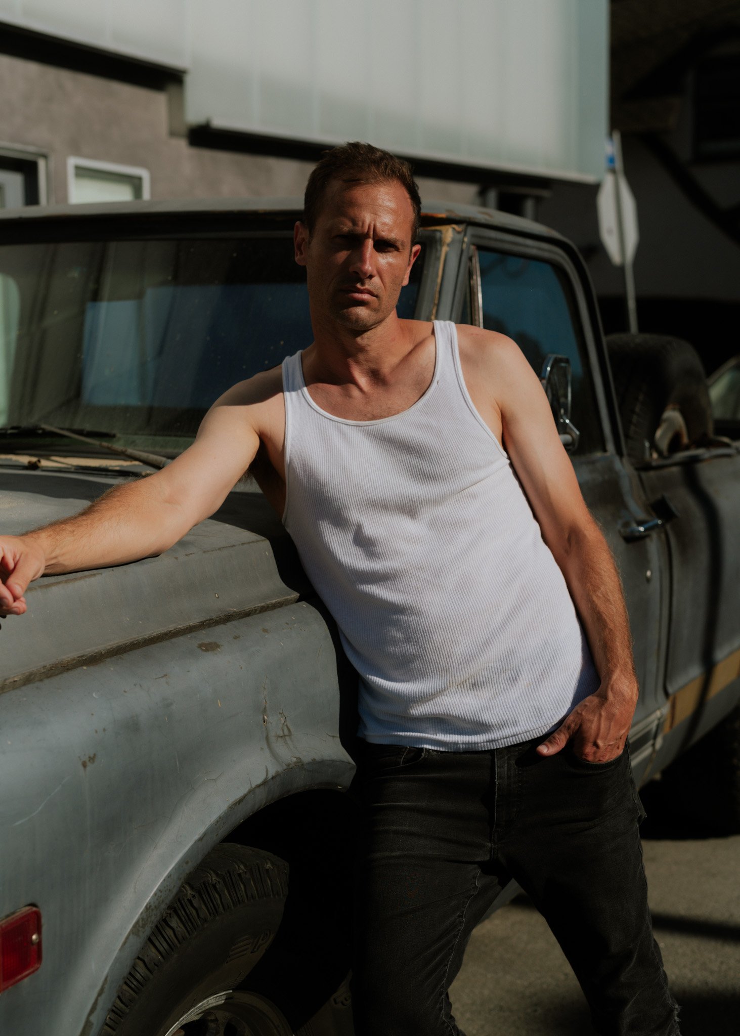A man in a white tank top and dark jeans posing beside an old, weathered pickup truck.