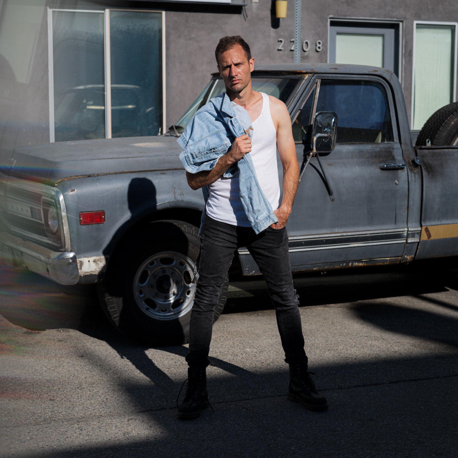 A man standing next to an old gray pickup truck, wearing a white tank top and a distressed denim jacket over his shoulder, with black pants and boots, in a parking lot outside a modern building.