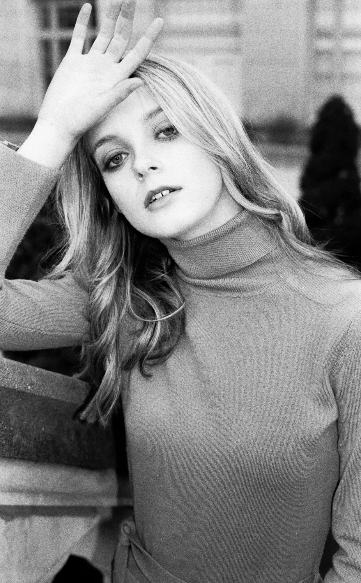 A woman with long, wavy hair is resting her hand on her forehead while looking into the camera. She is wearing a turtleneck sweater and appears to be sitting outdoors.