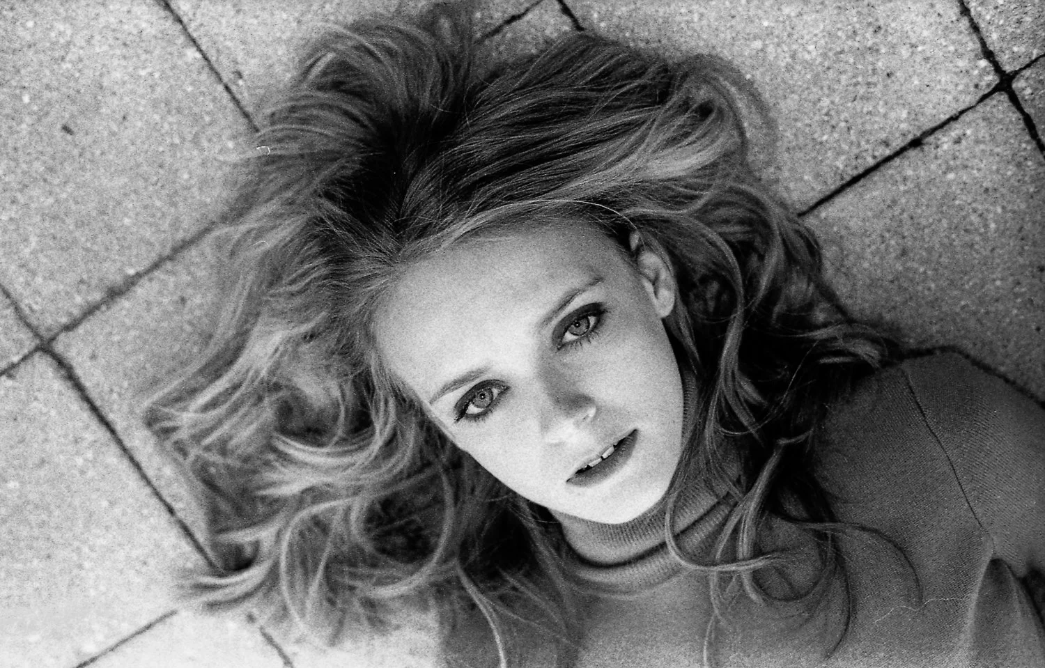 A woman with long, wavy hair lying on a stone or concrete surface, looking at the camera with an intense expression.