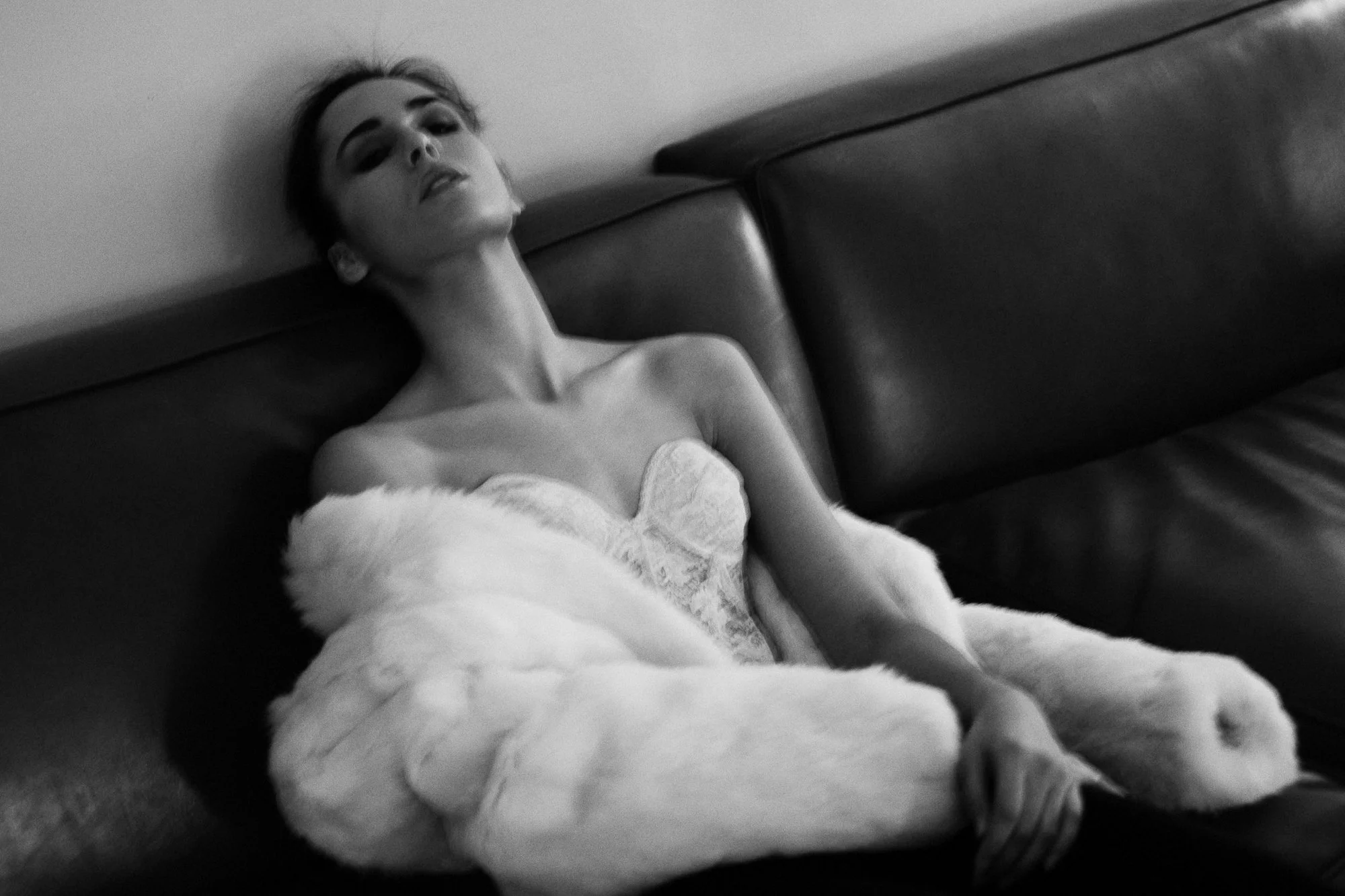 A black and white photo of a woman with her head tilted back, sitting on a couch, wrapped in a fluffy blanket, looking relaxed.