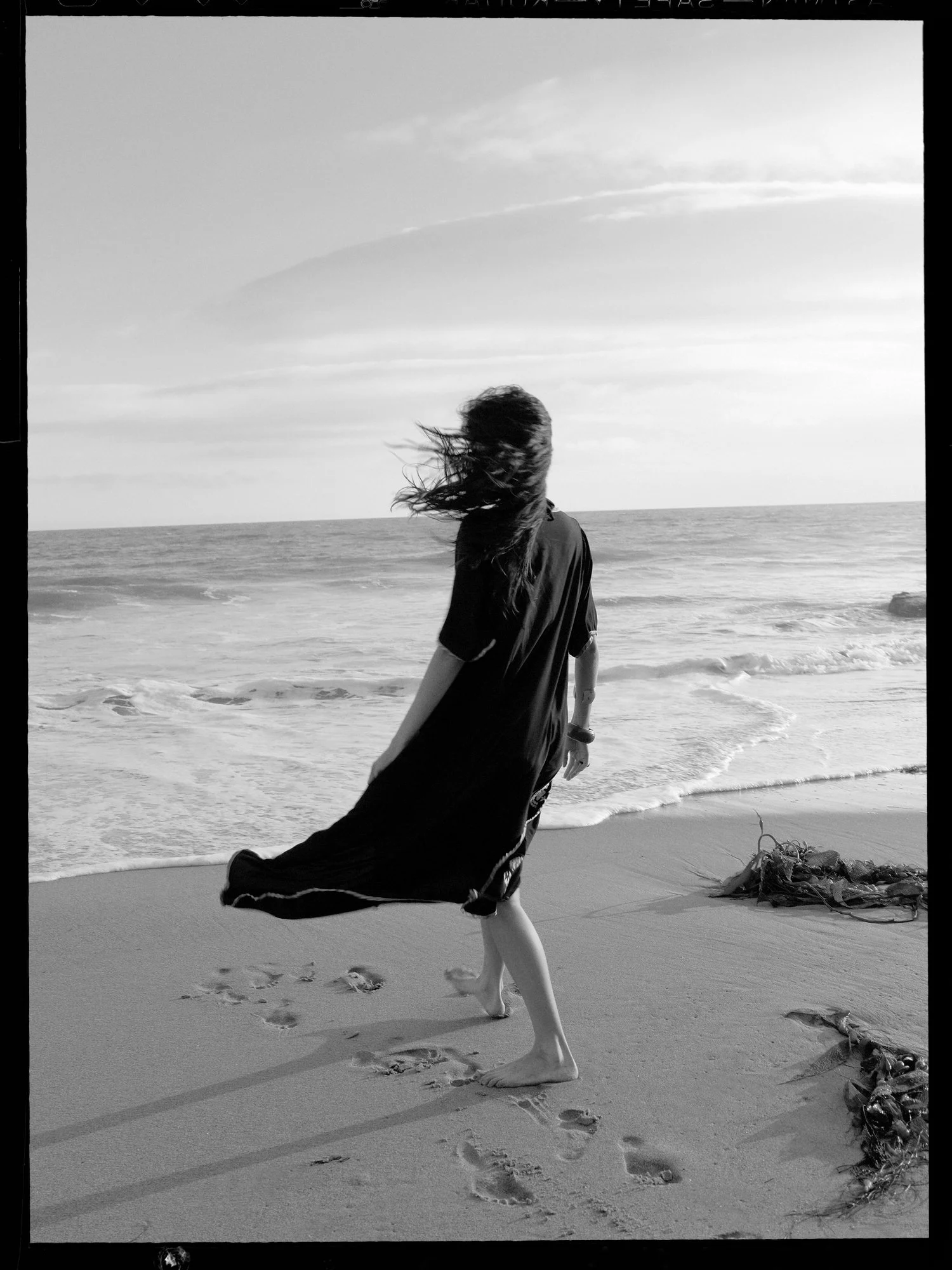 A woman with long hair standing on a beach, facing the ocean with her hair blowing in the wind. She is wearing a dark dress or cover-up, and there are footprints in the sand behind her. The sky is partly cloudy and the tide is coming in.