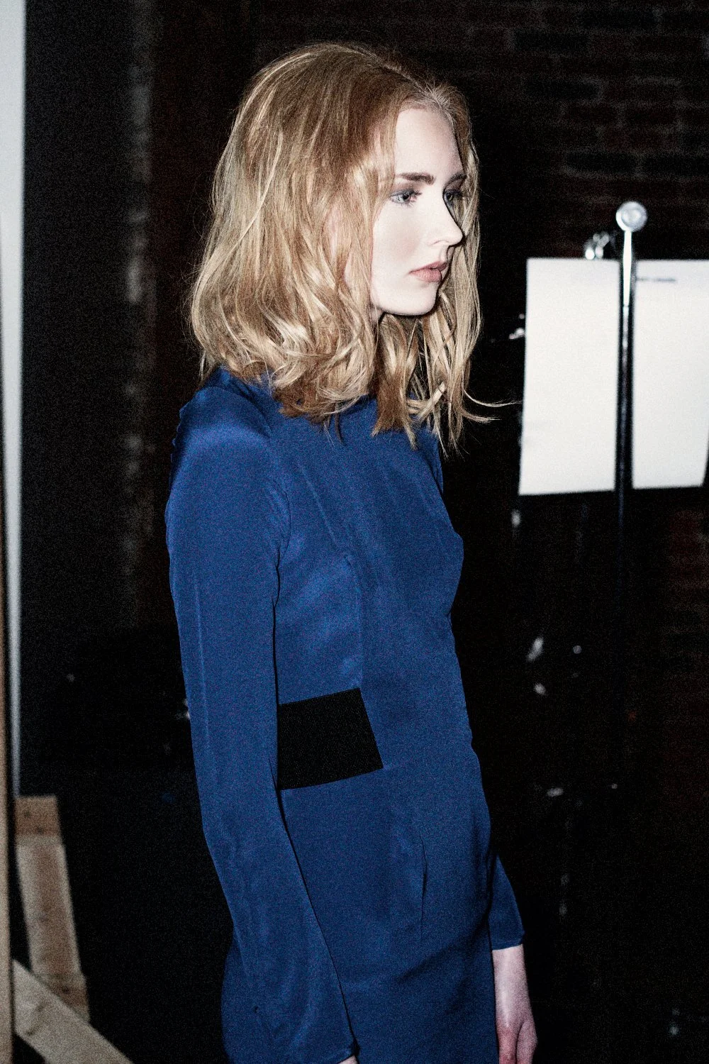 A woman with auburn hair is standing in profile, wearing a blue long-sleeve dress or top, against a dark background.