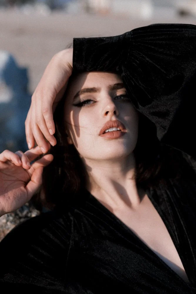 Close-up of a woman posing outdoors, with one hand on her head and the other near her face, wearing a black outfit and looking into the camera.