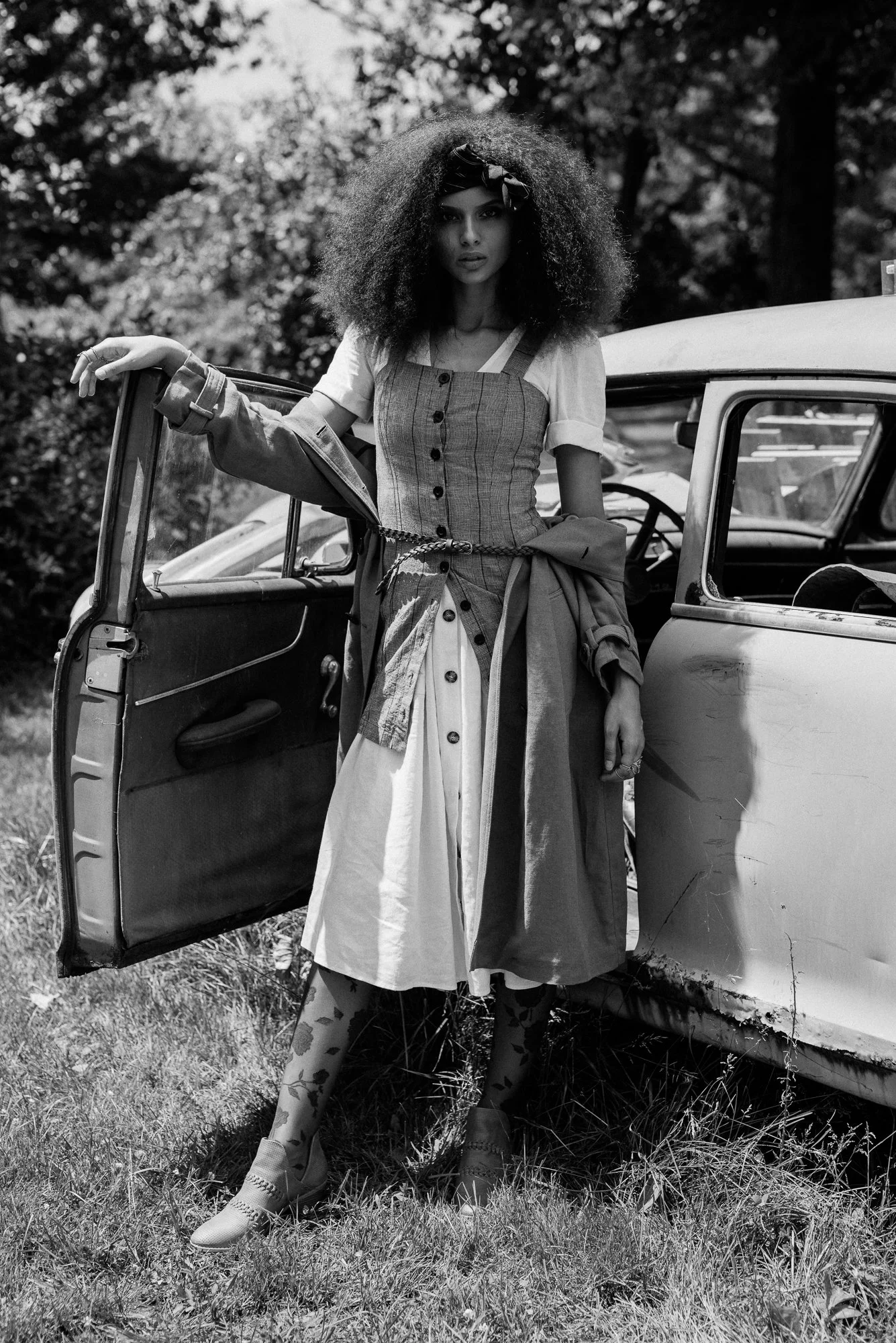 A woman with big, curly hair, wearing a plaid dress layered over a T-shirt, standing beside an open vintage car door in a grassy outdoor setting. The photo is in black and white.