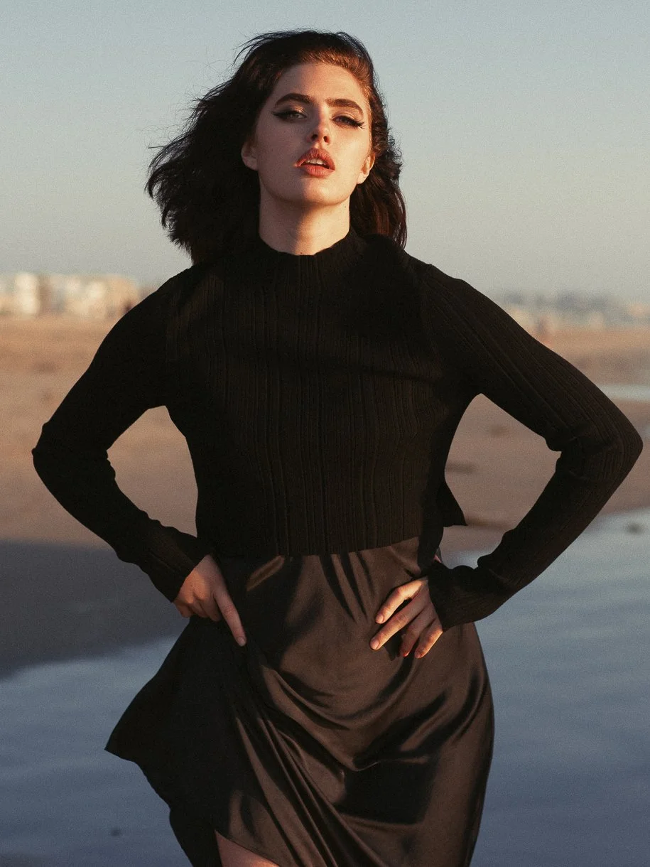 A woman with dark hair wearing a black long-sleeve top and black skirt, standing outdoors at sunset near a beach.