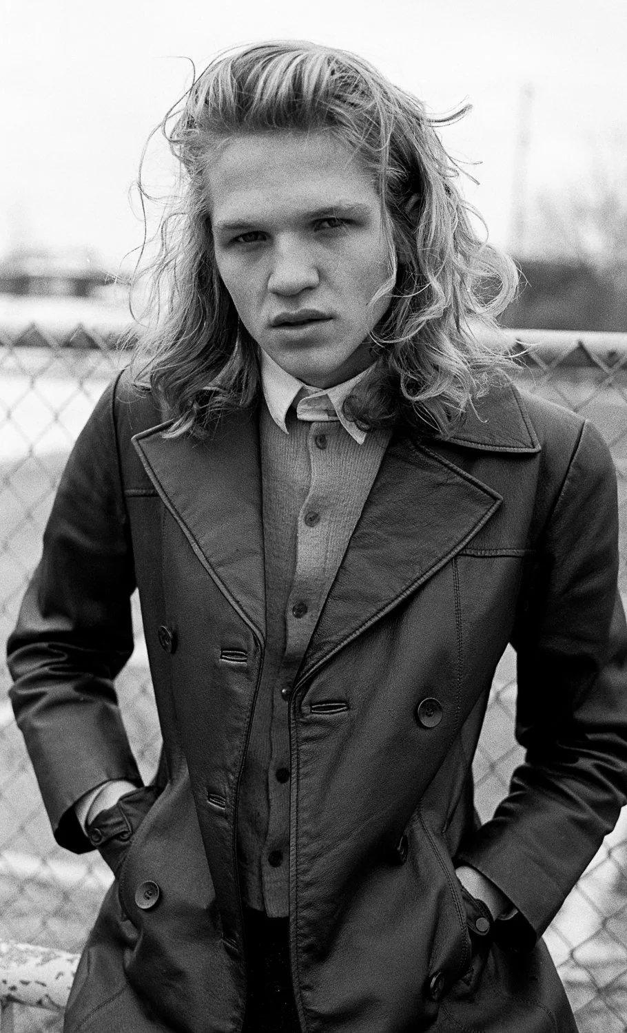 Black and white photo of a woman with wavy hair, wearing a leather coat over a shirt, standing outdoors in front of a chain-link fence.