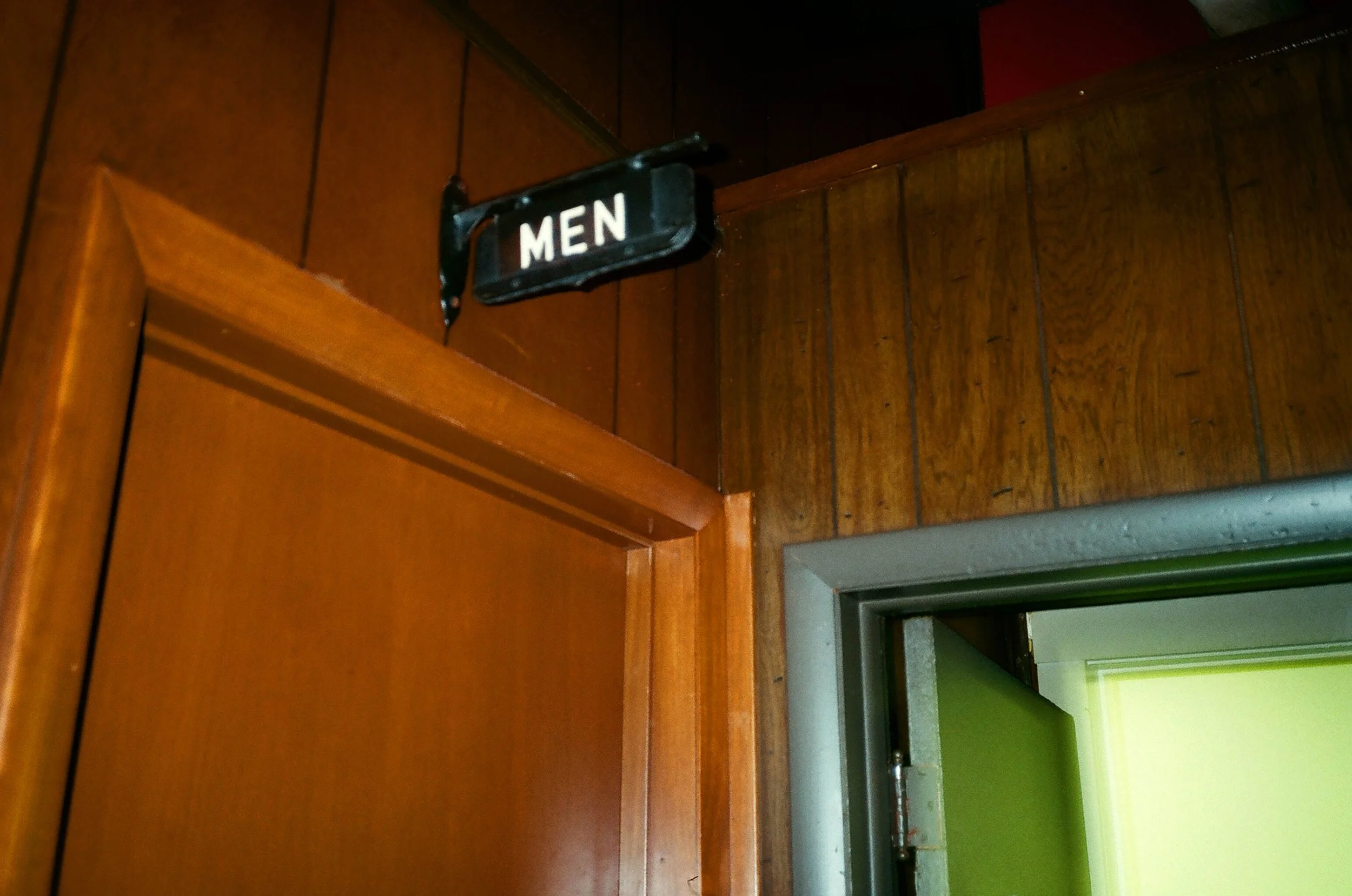 A wooden bathroom door with a black sign that says 'MEN' hanging on the top part of the door. The door is part of a wall with similar wooden paneling. To the right of the door, there is a restroom area with brightly painted walls and details, includi