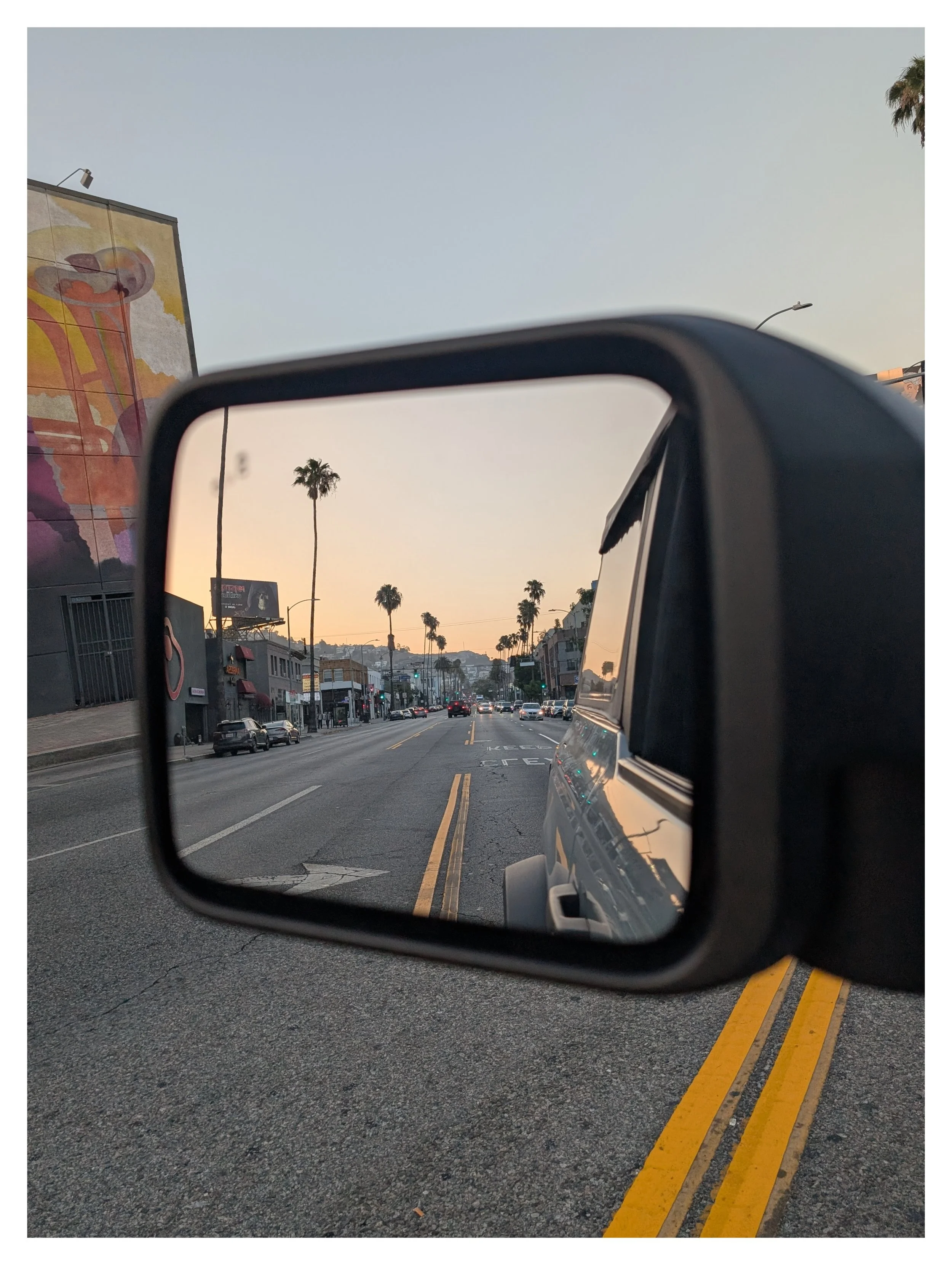 Photo taken from the side mirror of a vehicle showing a city street with parked cars, palm trees, buildings, and a twilight sky.