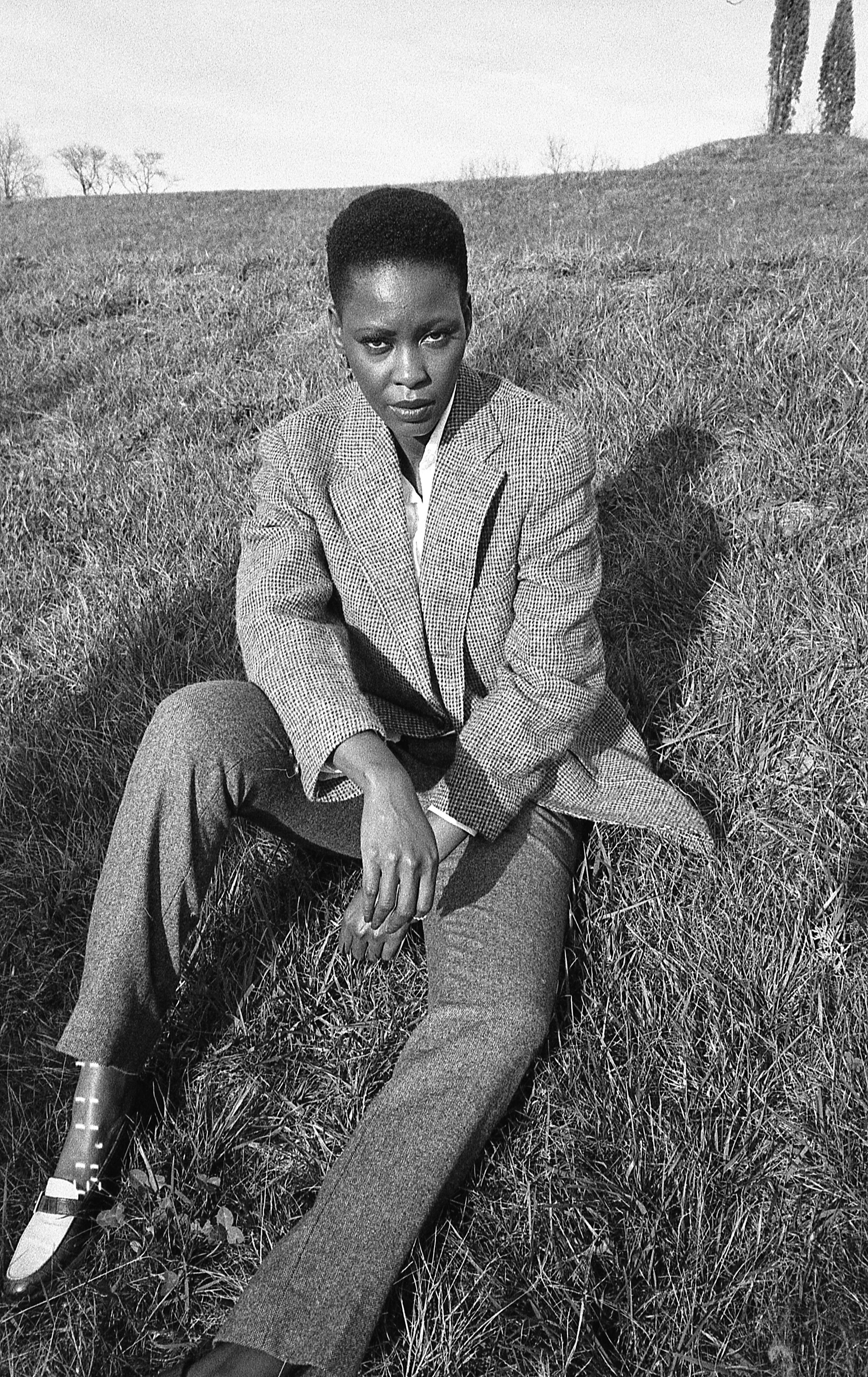 Black and white photo of a woman sitting on grass in an open field, wearing a checkered blazer and trousers, with trees and a hill in the background.