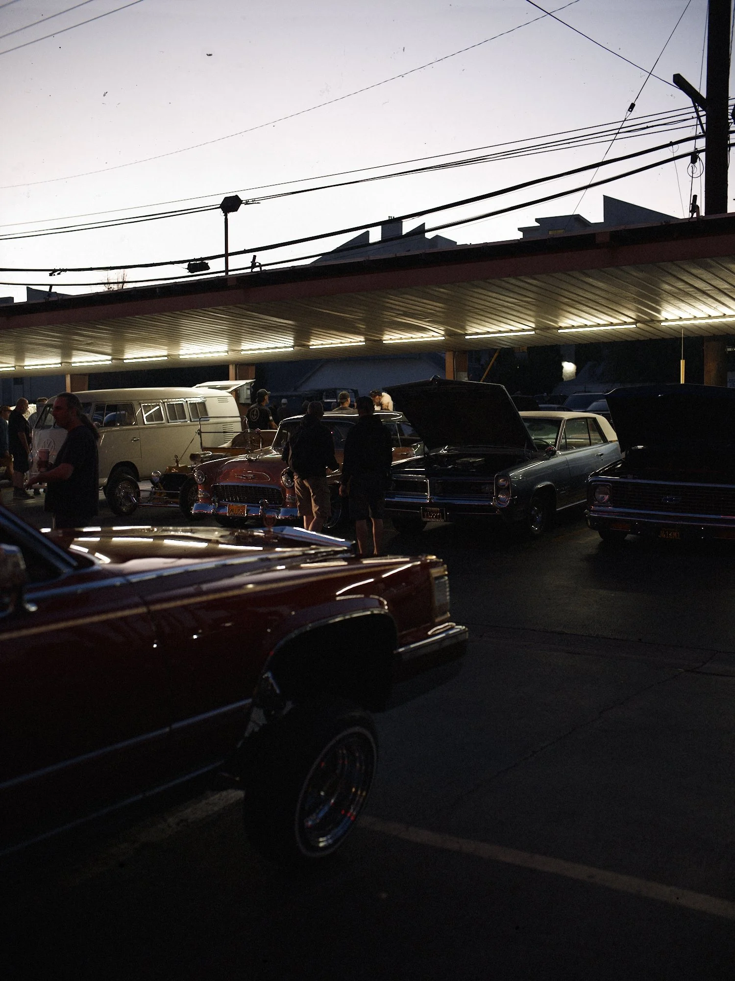 Nighttime parking lot filled with vintage cars and people, illuminated by overhead lights, with a building and power lines in the background.