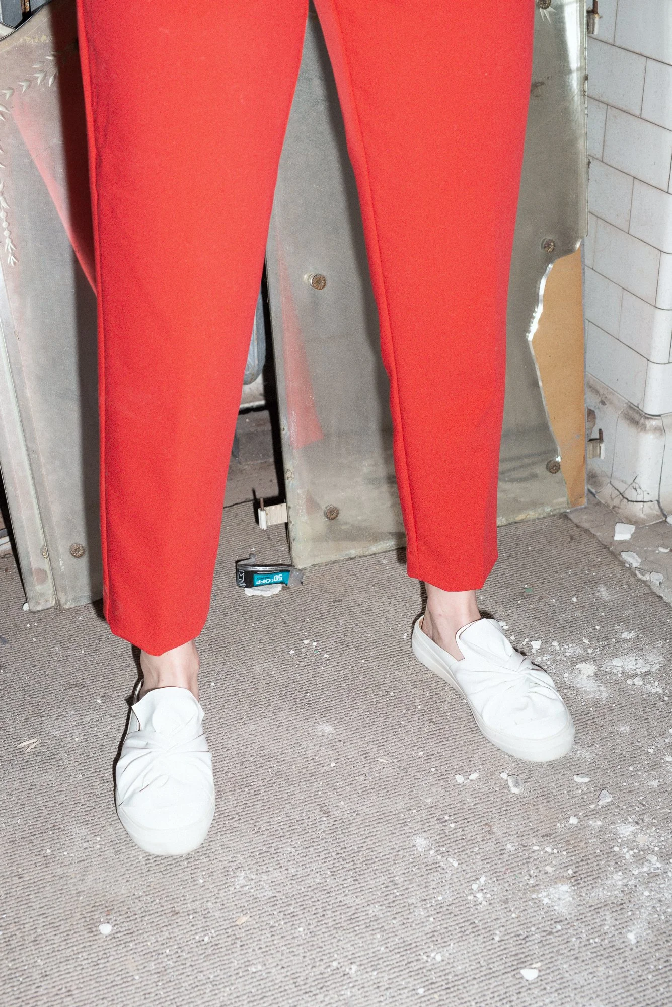 Person wearing red pants and white sneakers standing on a carpeted floor with scattered debris, behind metallic equipment and white brick wall.