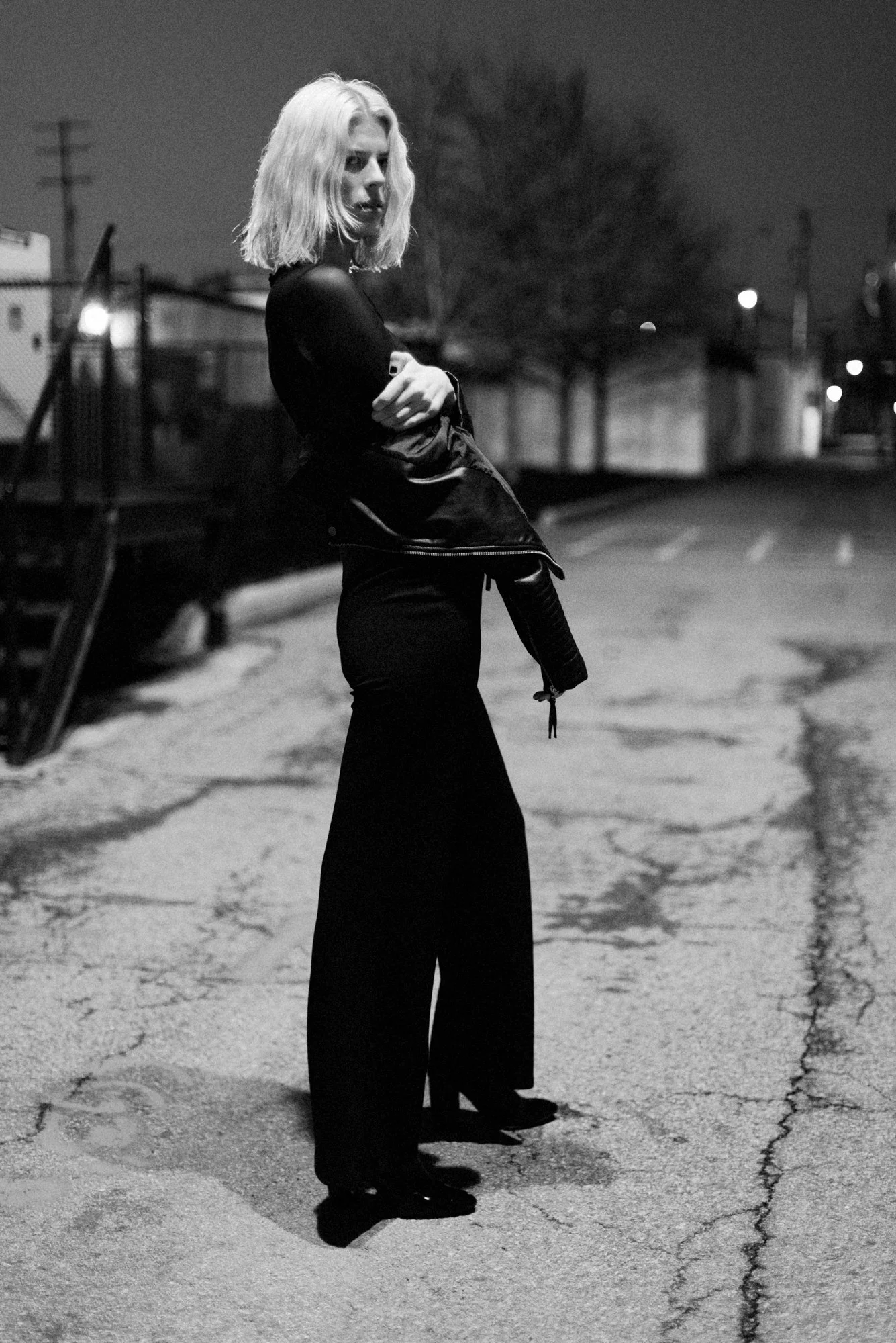 Black and white photo of a young woman with shoulder-length hair standing outdoors at night on a cracked pavement street. She is wearing a long skirt, a dark top, and holding a jacket, with her arms crossed. In the background, blurred leafless trees,