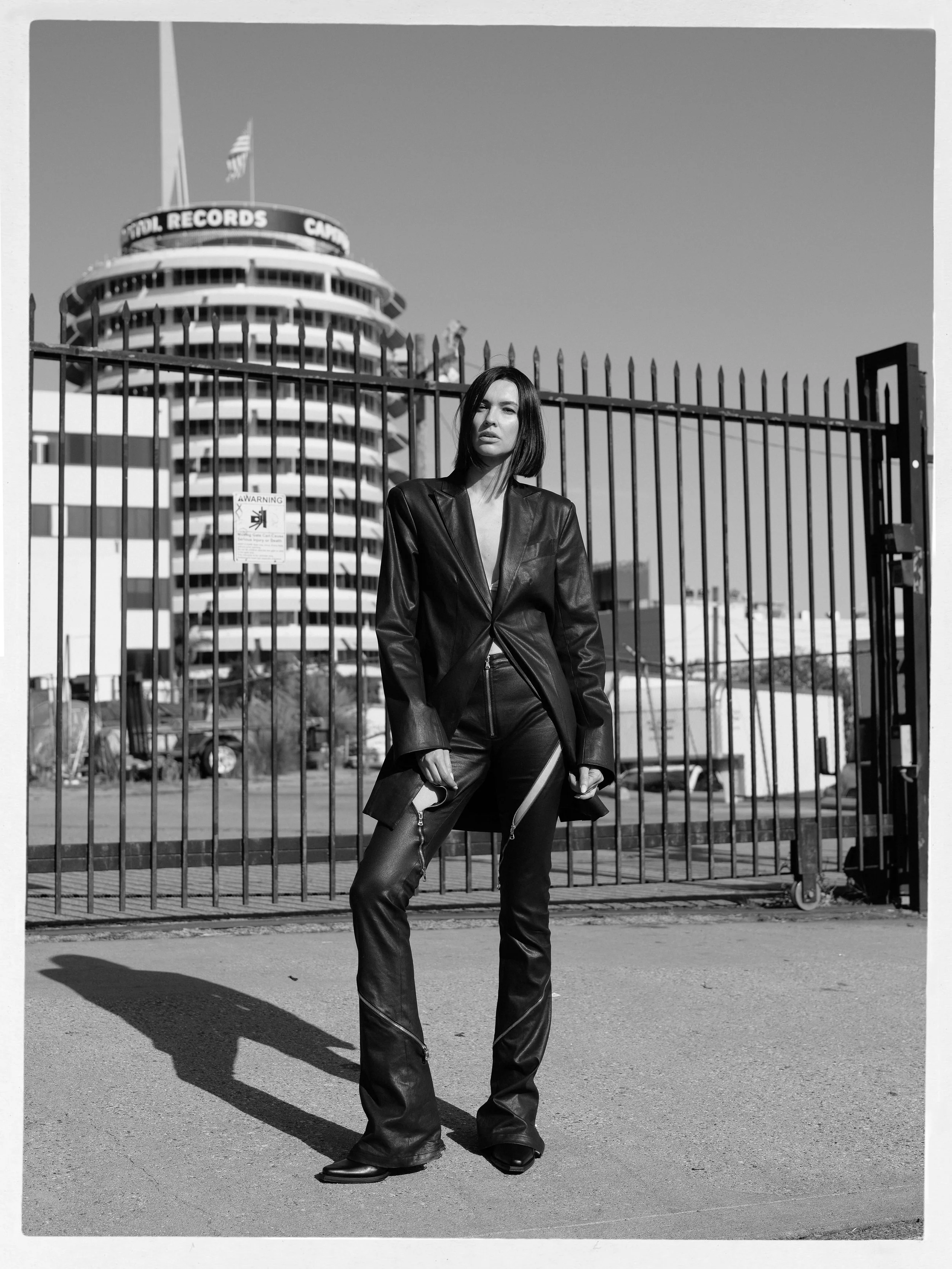 A woman in a leather jacket and leather pants standing outdoors in front of a metal fence. There is a modern multi-story building with a sign reading "STUDIO RECORDS" and an American flag behind the fence. The woman has dark hair and a serious expres