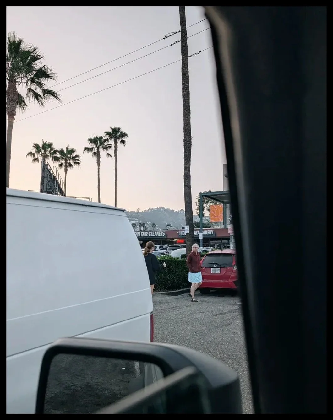 A scene in a parking lot with two women conversing near a red car, palm trees in the background, and storefront signs indicating a bakery and dry cleaners.