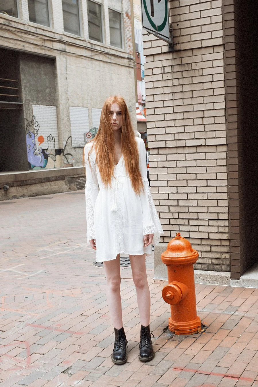 A young woman with long red hair standing on a brick sidewalk in an urban alleyway near a brick wall and fire hydrant.