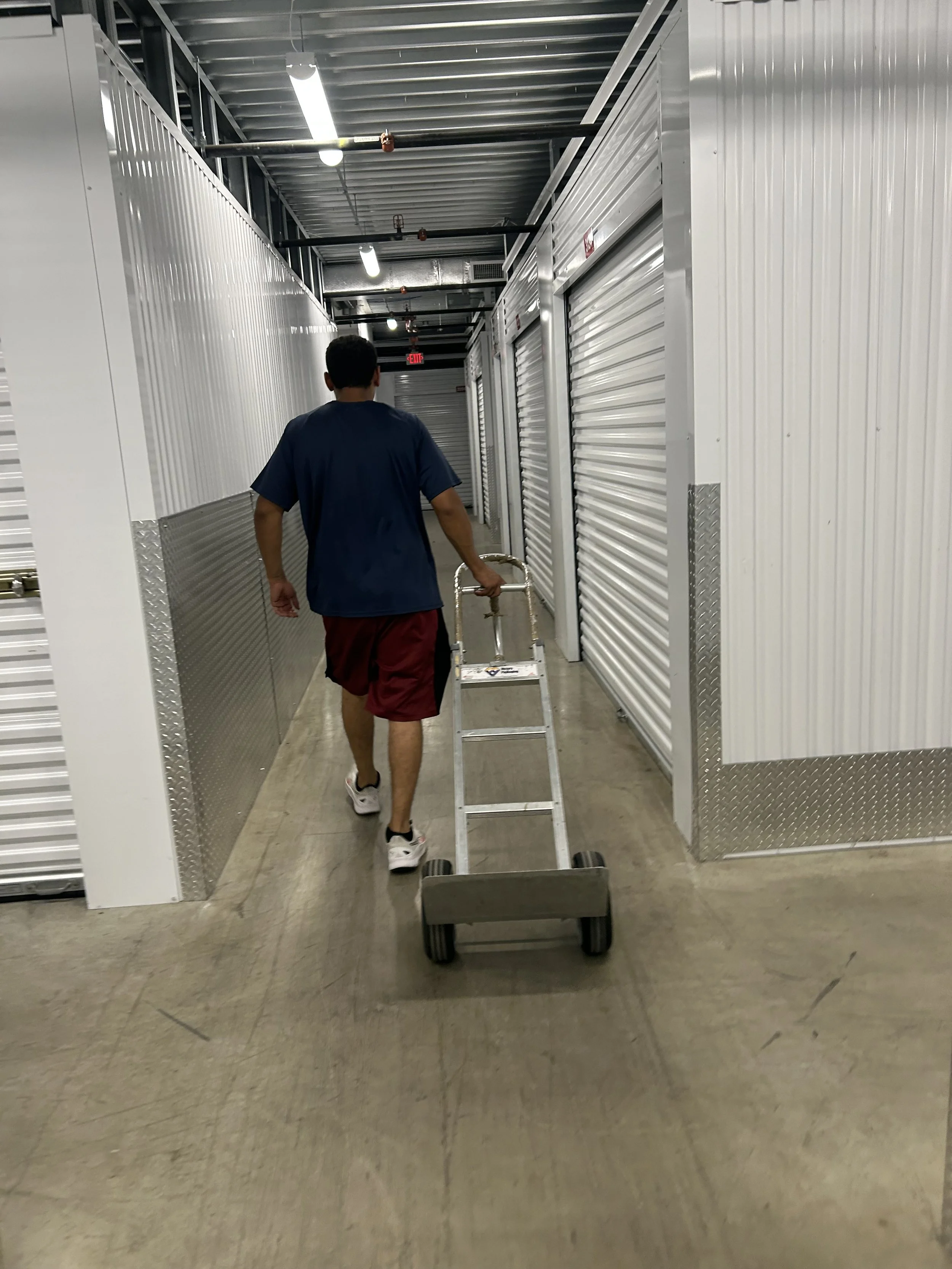 A person walking in an indoor self-storage facility hallway with white storage unit doors on the sides, a rolling hand truck in front, and overhead fluorescent lighting.