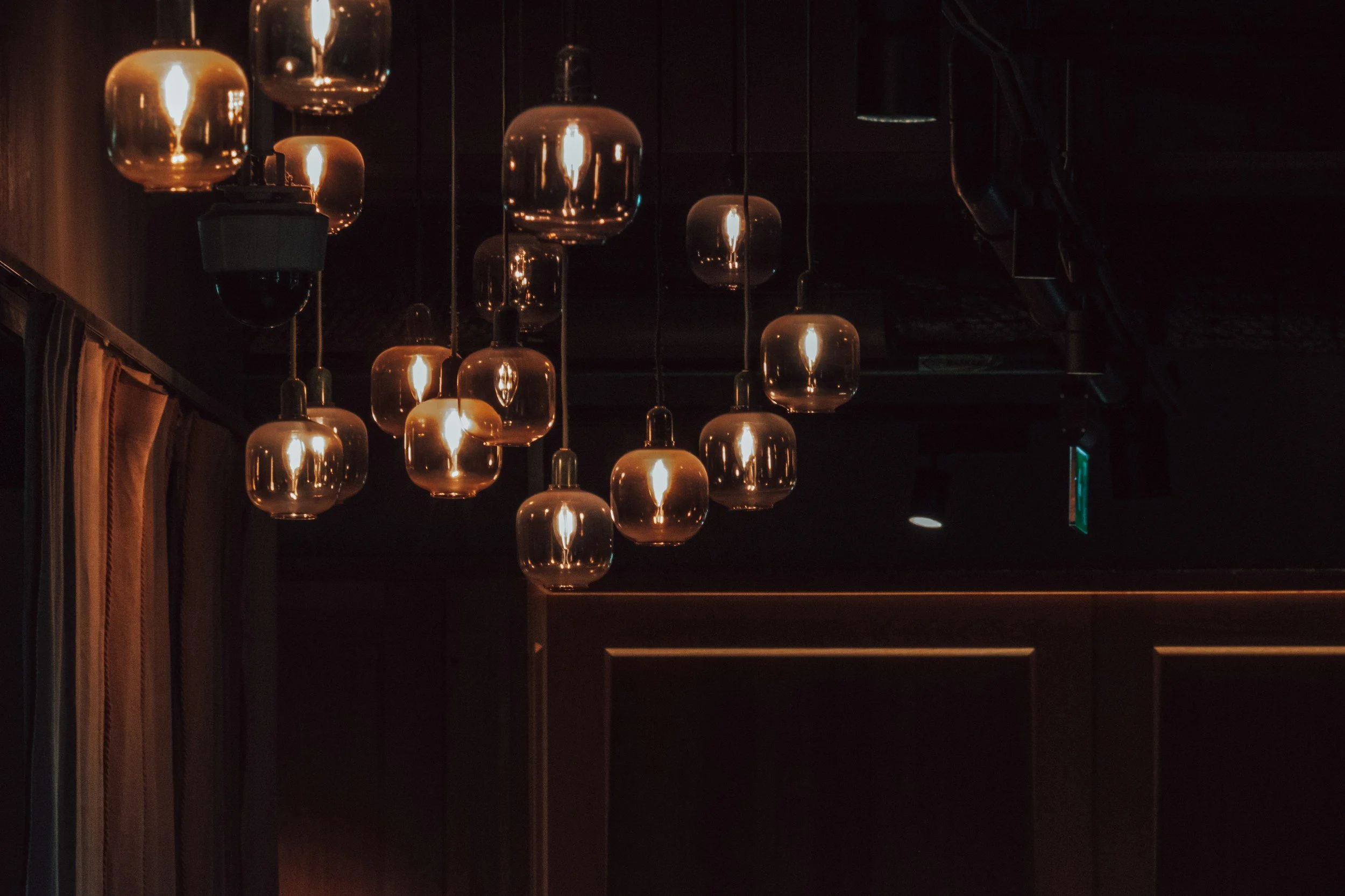 A cluster of vintage-style pendant lights with amber glass globes casting a warm, atmospheric glow in a dimly lit, moody room. The lights hang at varied heights above dark wood wall panels and elegant fabric drapery, evoking an intimate supper club.