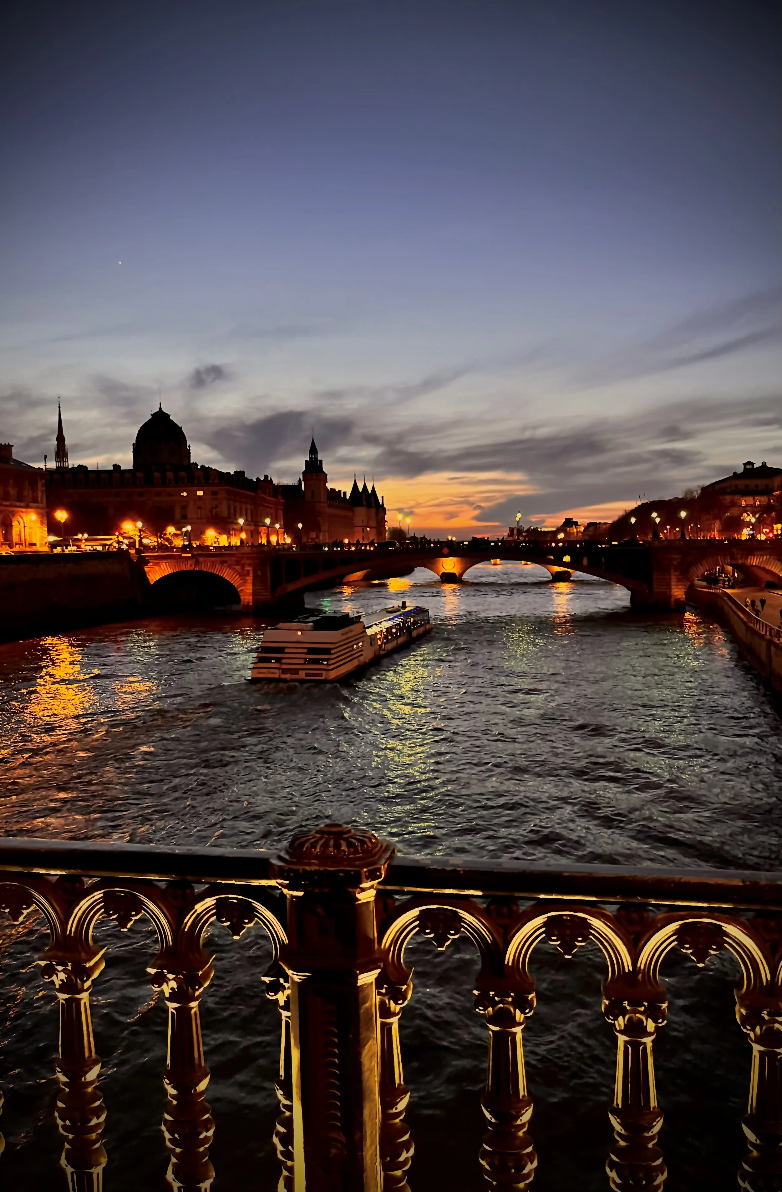 View of a bridge over a river at sunset with city buildings and spires in the background, illuminated by streetlights, and a boat on the water.