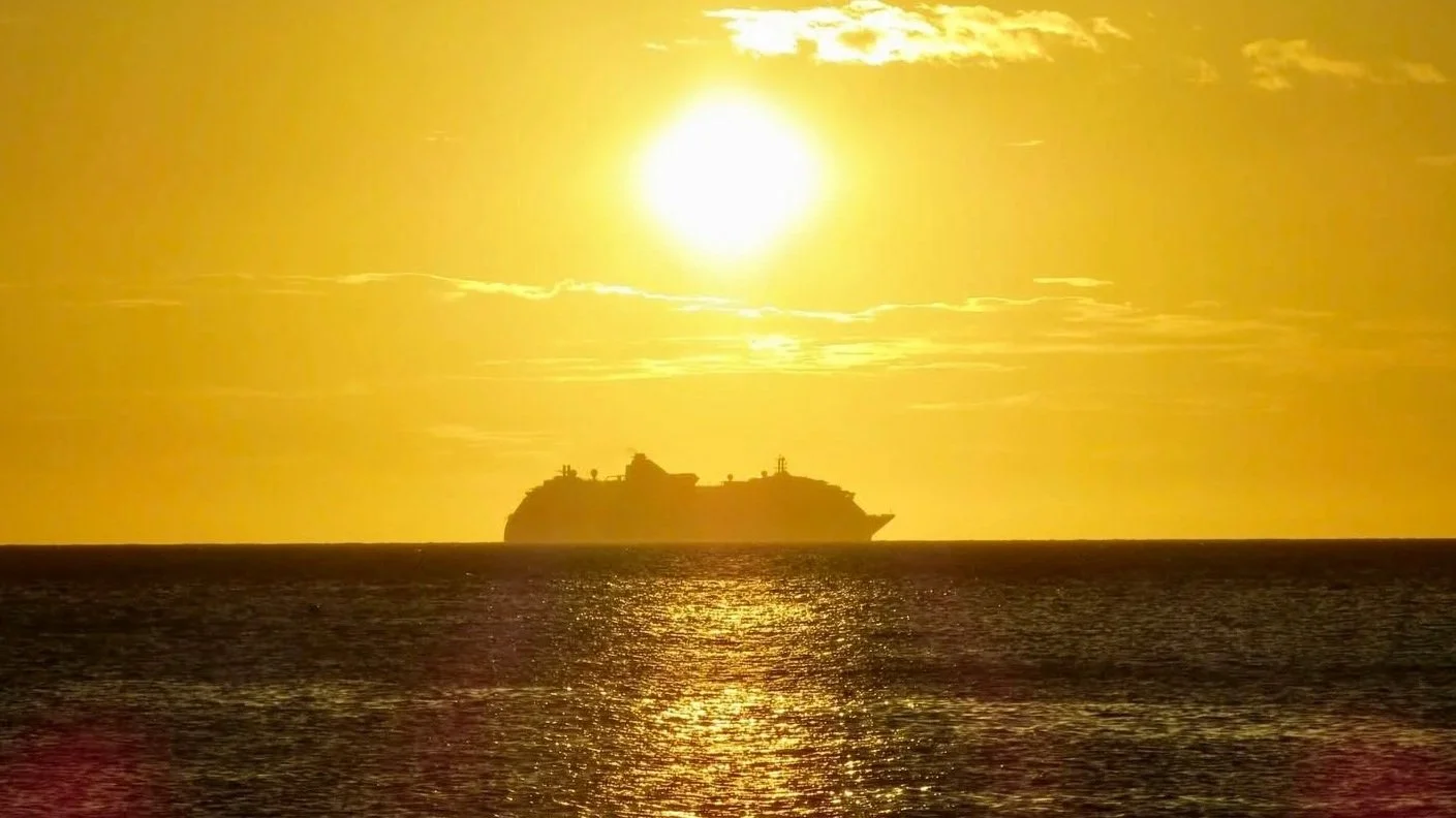 Silhouette of a large ship sailing on the ocean during sunset with a bright yellow sky and shimmering water.