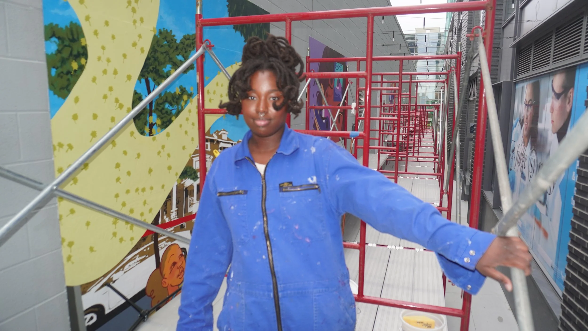A person with dreadlocks standing on a scaffold in front of a colorful mural, wearing a blue jacket covered in paint splatters, holding onto scaffolding bars with one hand.