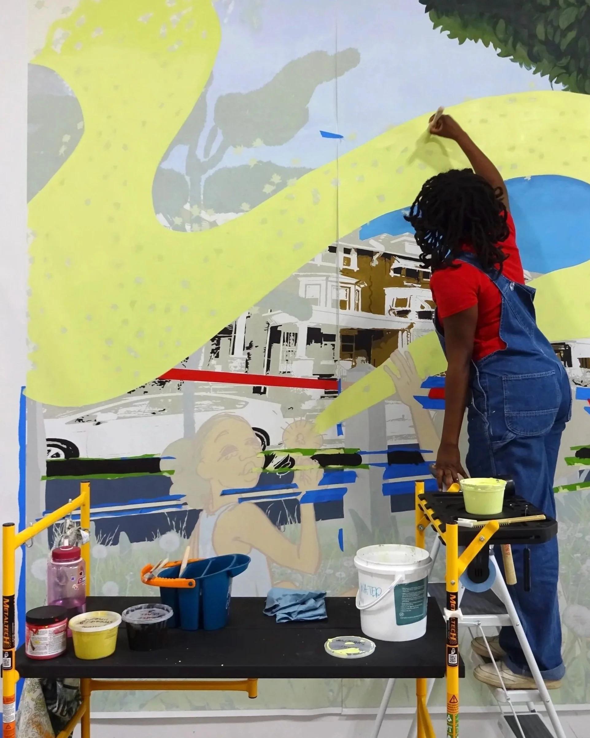 A person painting a mural on a wall with various paint supplies and brushes on a table and utility tower.