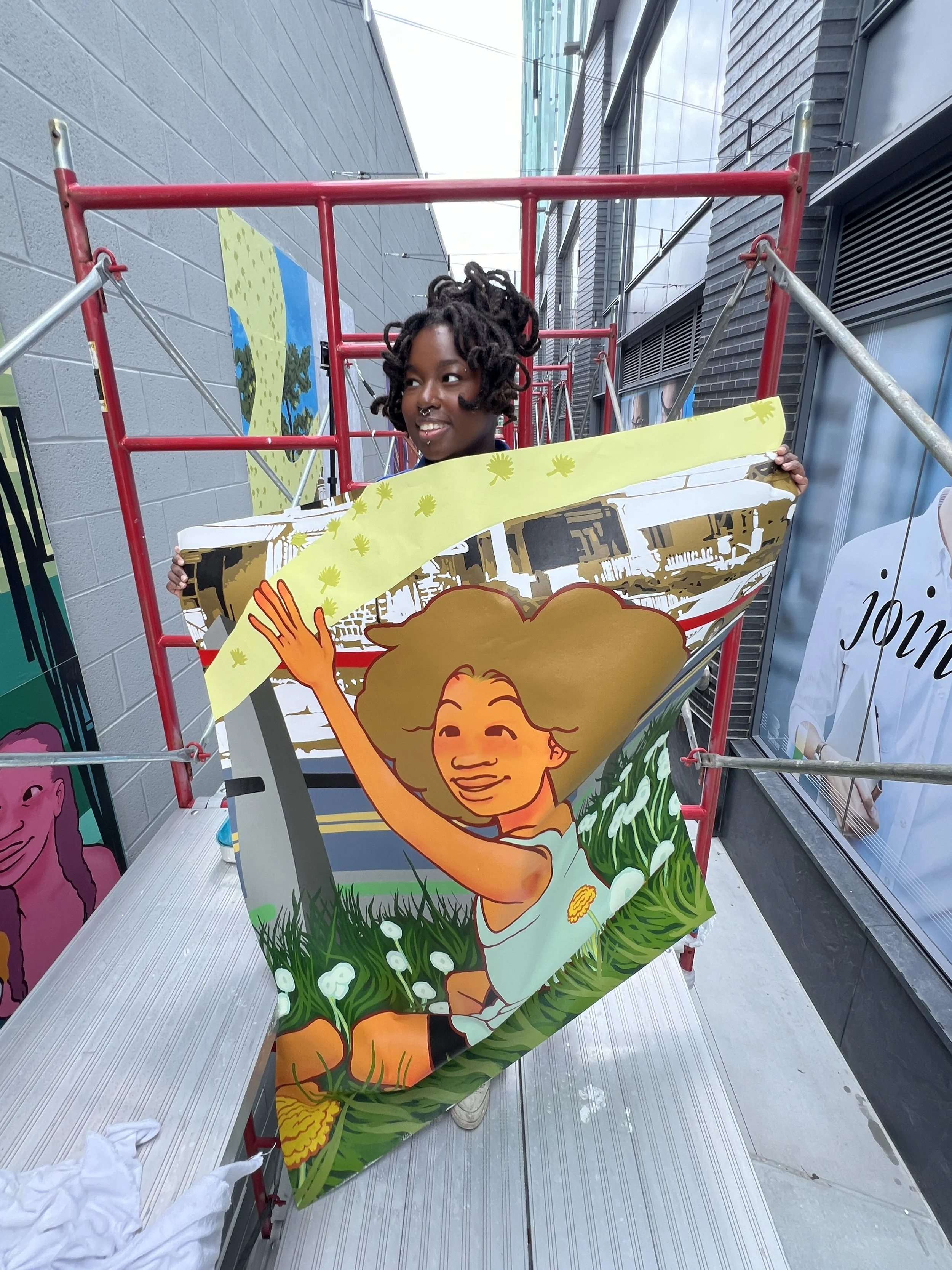 A person with dark skin and dreadlocks smiling while holding up a colorful illustrated sign or piece of artwork featuring a smiling character with curly hair, surrounded by green grass and white flowers, positioned on a metal platform outdoors with city buildings in the background.